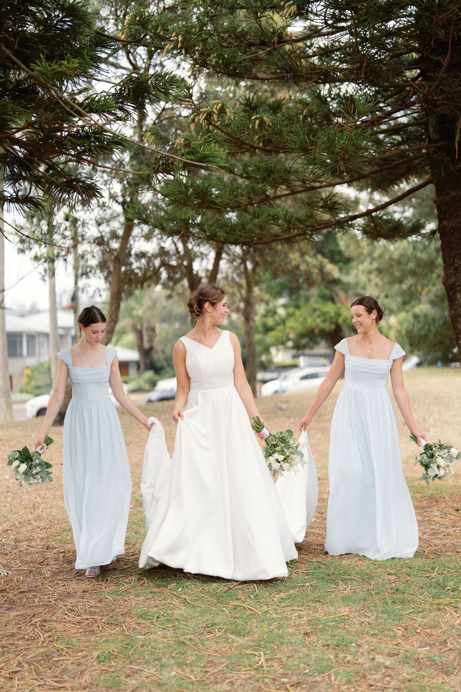 A group in white dresses walks together through a pine forest setting during an outdoor photo session.