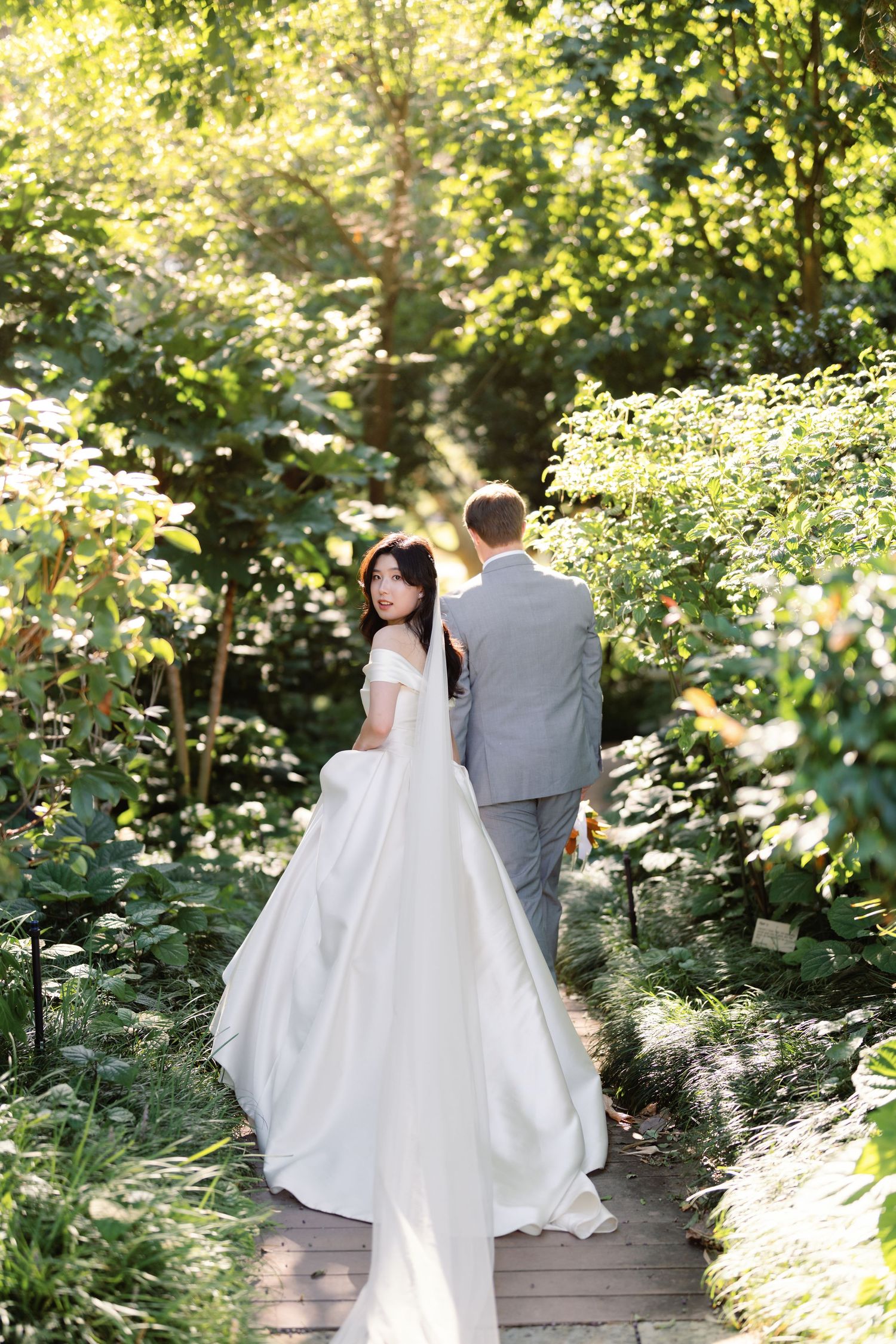 Wedding couple walks together on garden pathway surrounded by lush greenery.
