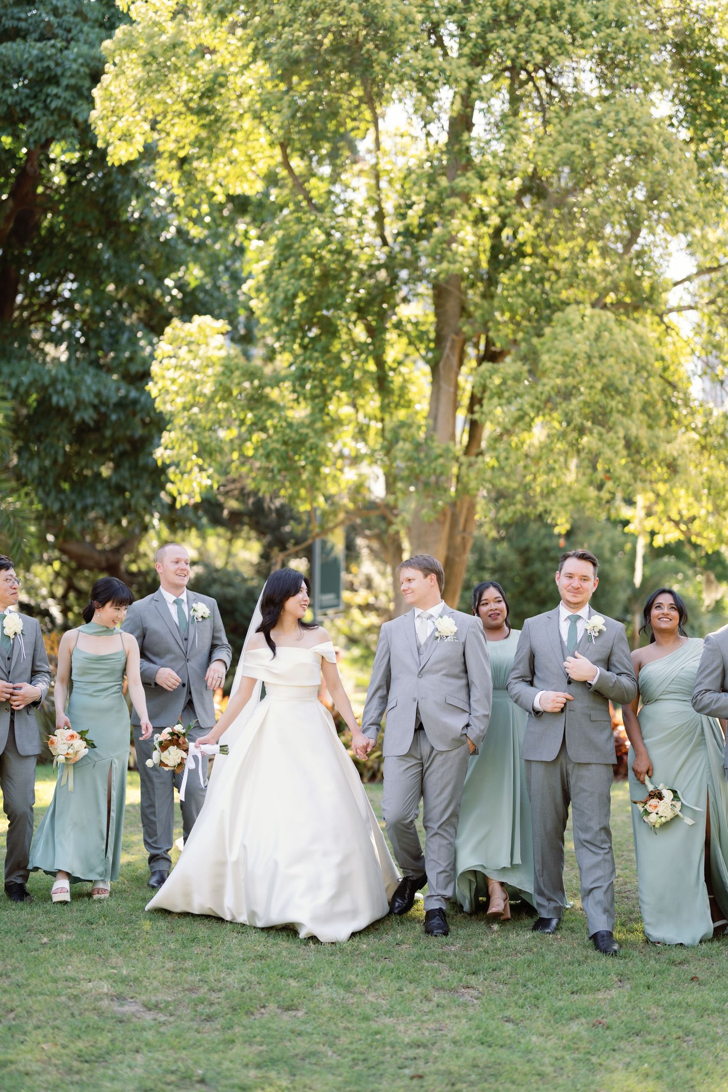 Wedding party in sage green and gray attire poses outdoors under sunlit trees.