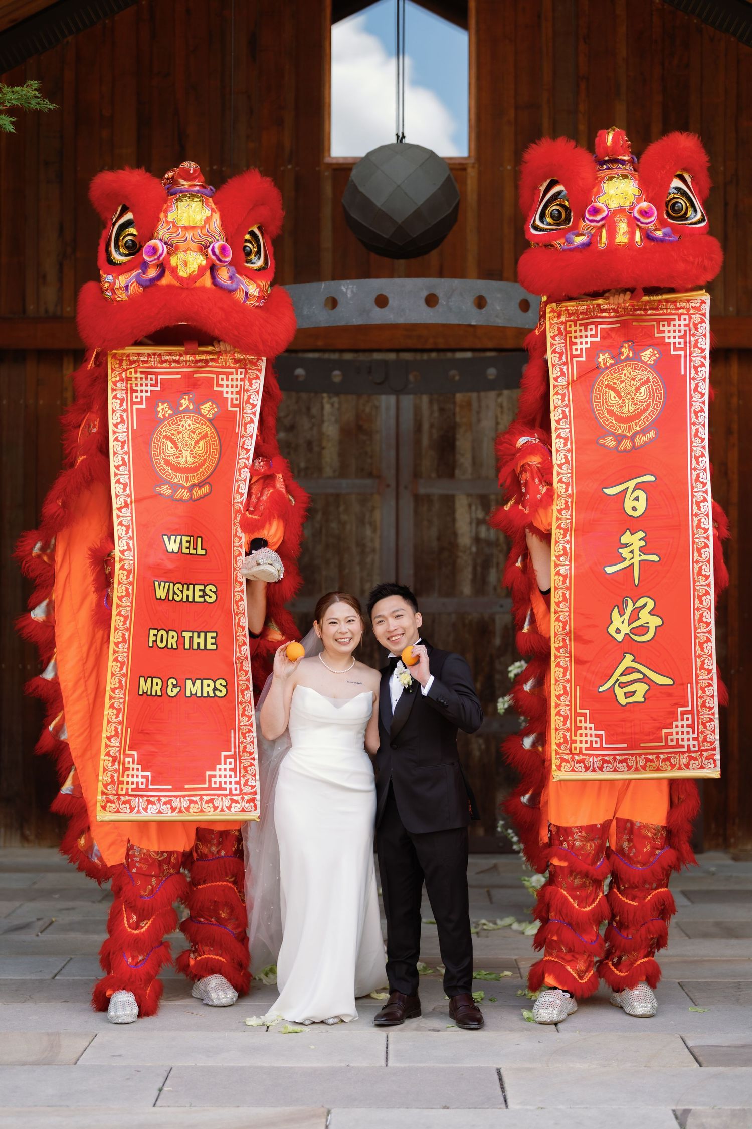 Traditional Chinese wedding banners and lion dance performers pose against wooden barn backdrop.