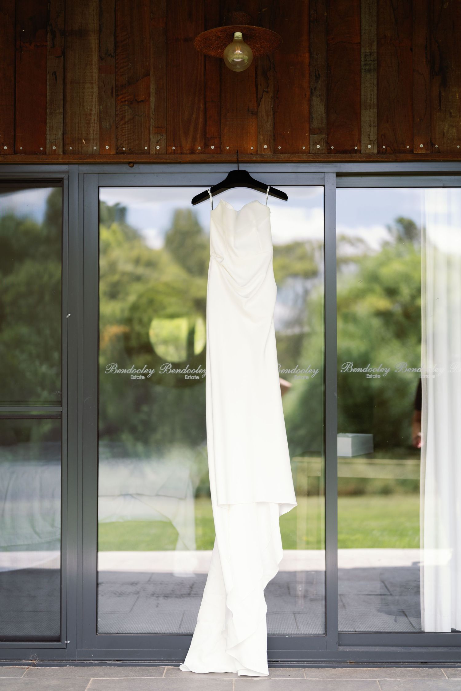White wedding dress hangs displayed against wooden paneled windows in natural light.
