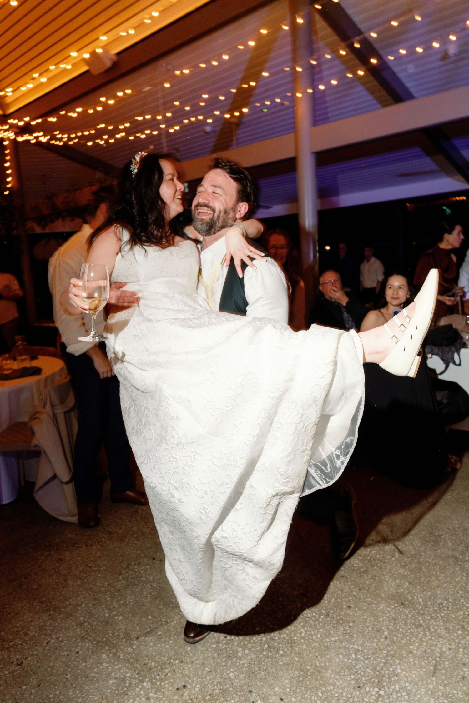 A joyful wedding celebration under twinkling string lights as a couple shares a romantic dance with the bride being lifted.