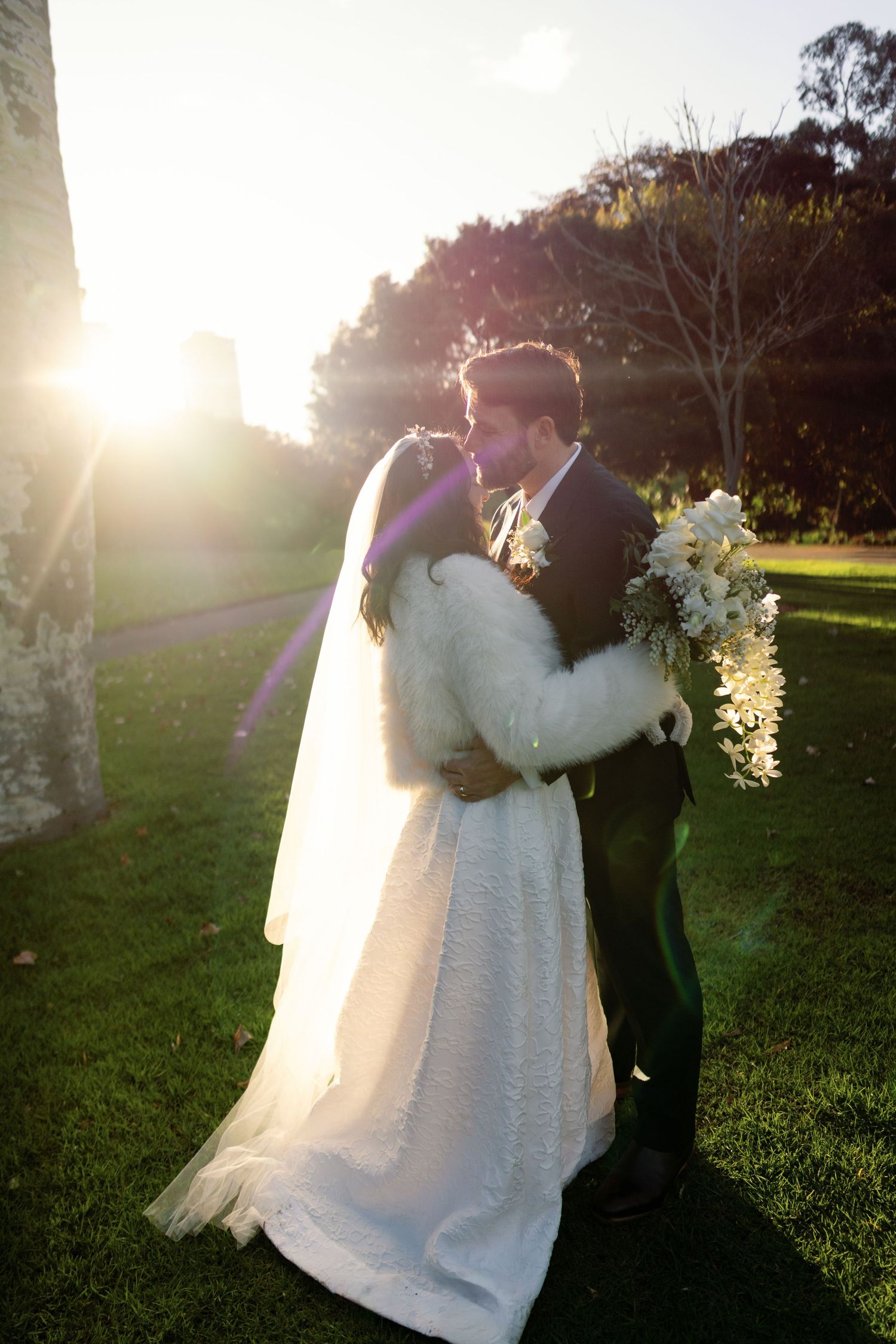 Bride and groom share a kiss at sunset in garden, bride holding white orchid bouquet.