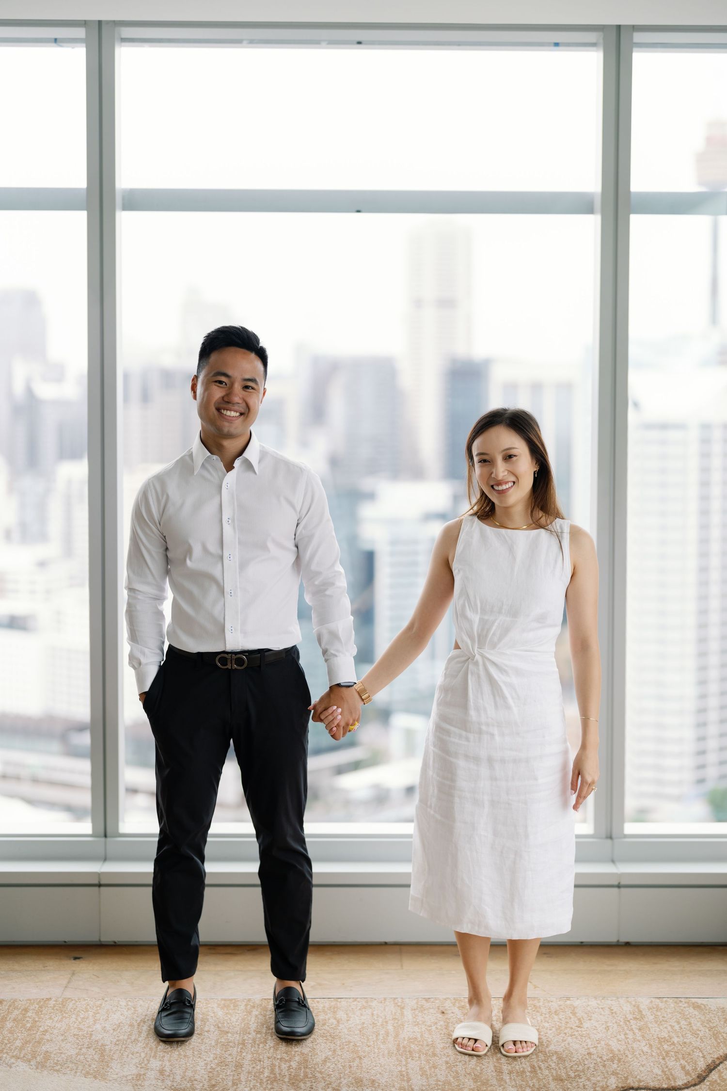 A couple in white attire standing together against a bright window overlooking a city skyline.
