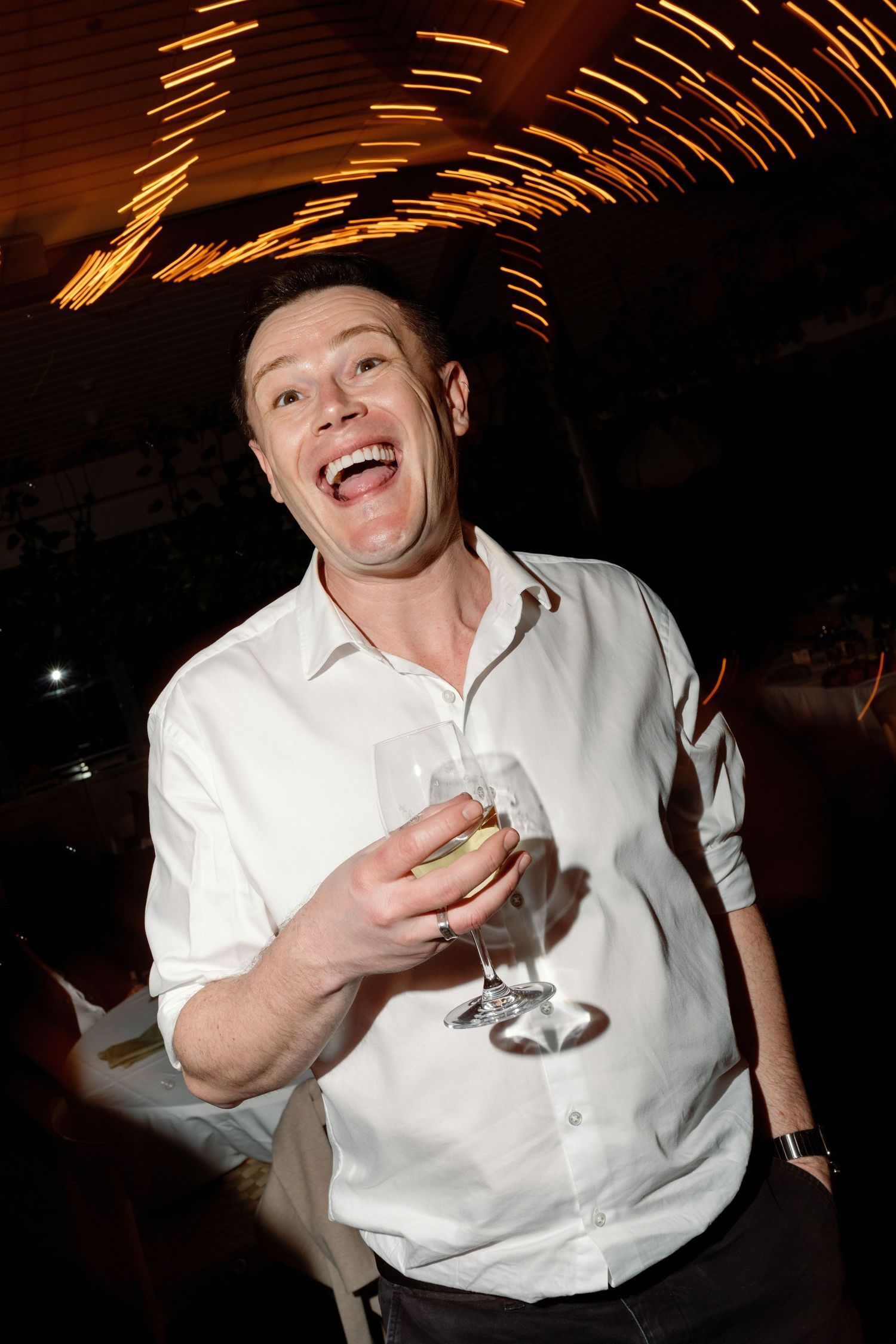 Joyful man in white shirt laughing and holding champagne glass at a lively wedding celebration.