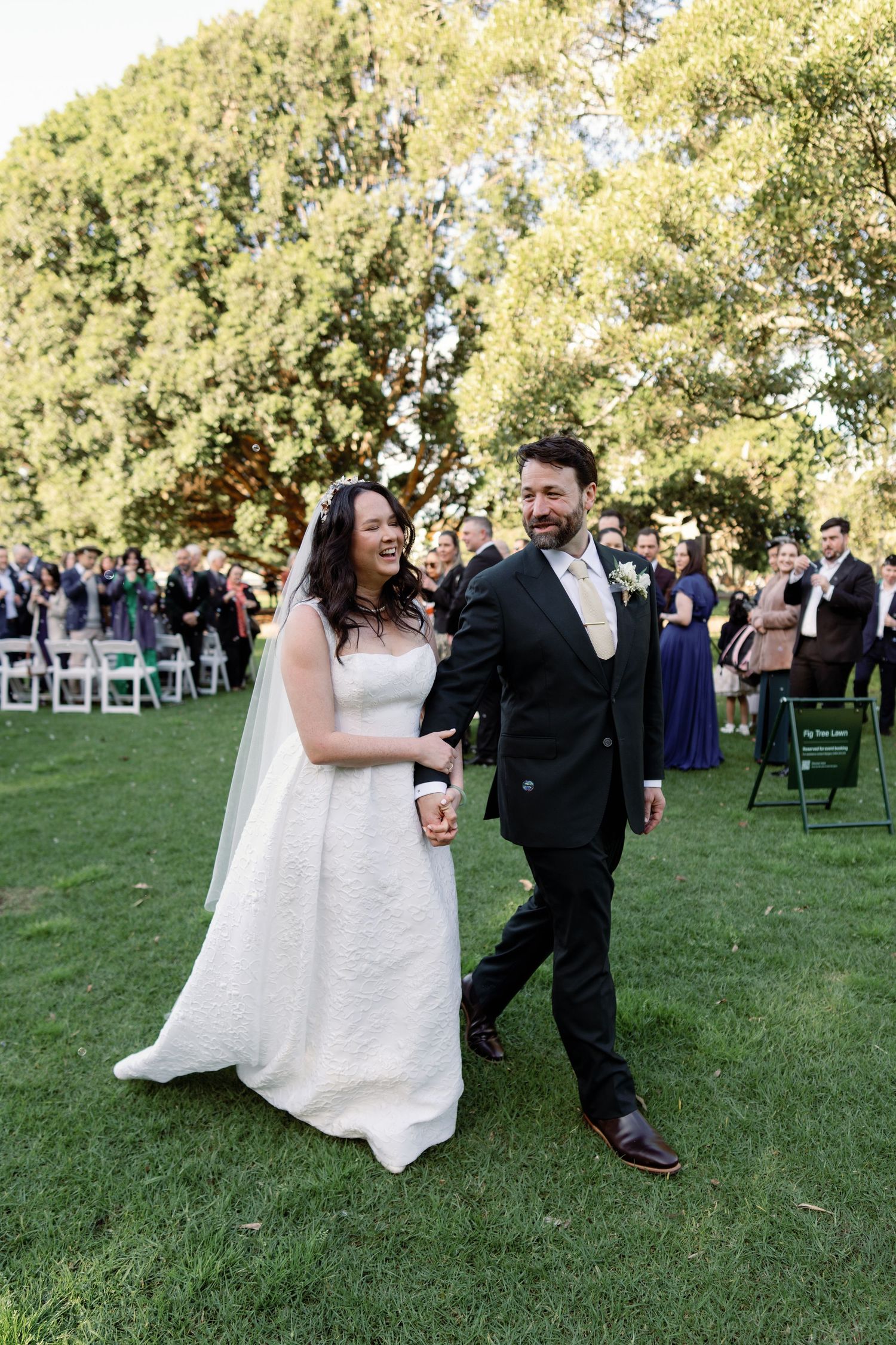 Newlyweds walk joyfully down outdoor aisle on grass surrounded by wedding guests.