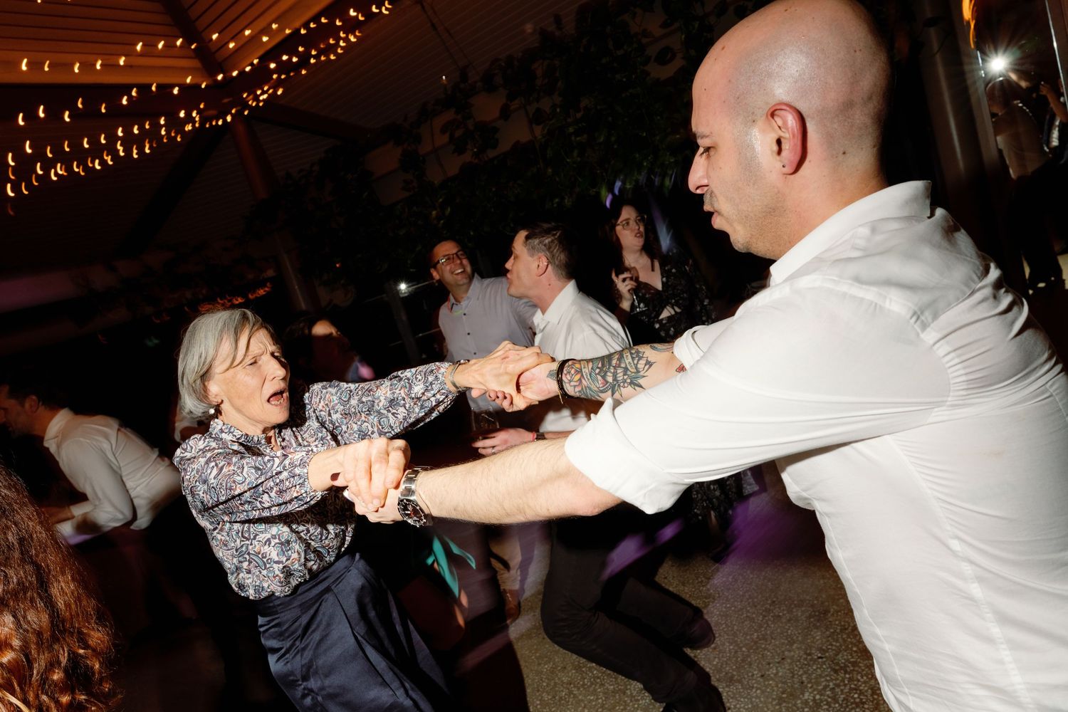 Elderly woman and bald tattooed man dancing energetically together at an indoor evening event.
