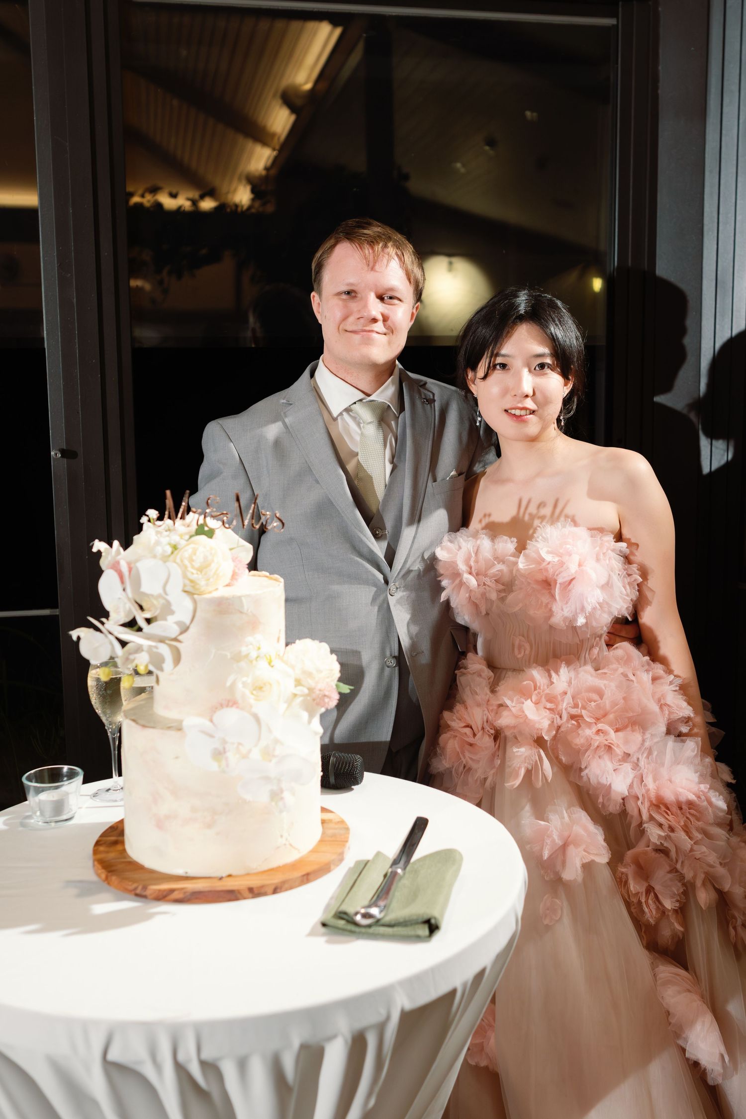 Wedding couple poses next to their elegant white tiered cake at reception.