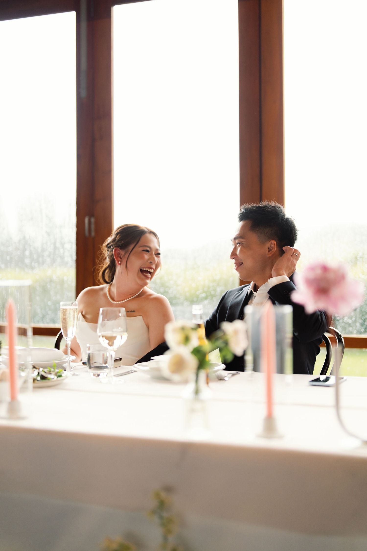Wedding guests sharing laughter at a reception table with large windows and natural light.