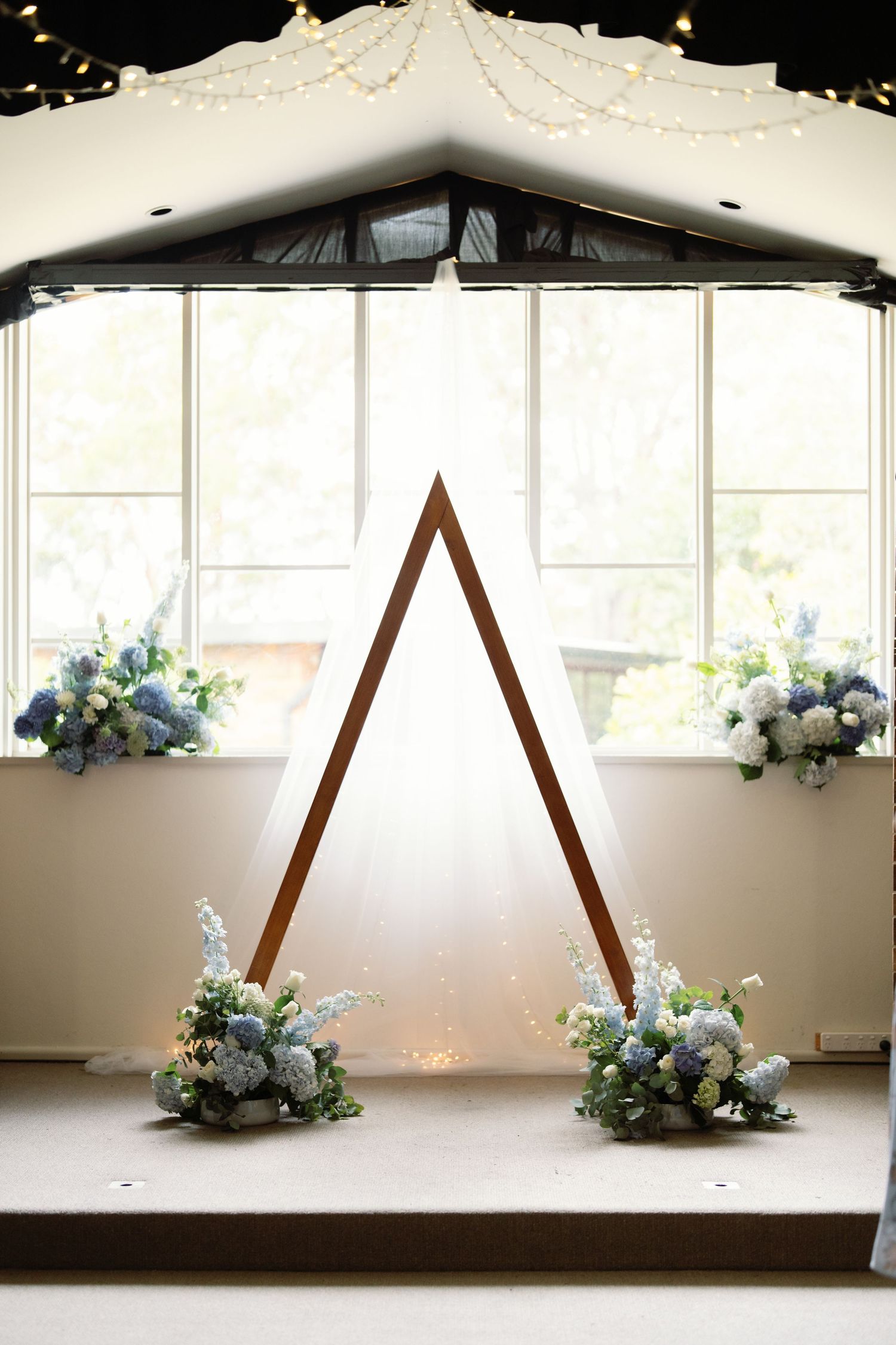 Wooden triangular wedding arch decorated with blue and white floral arrangements in a bright indoor venue.