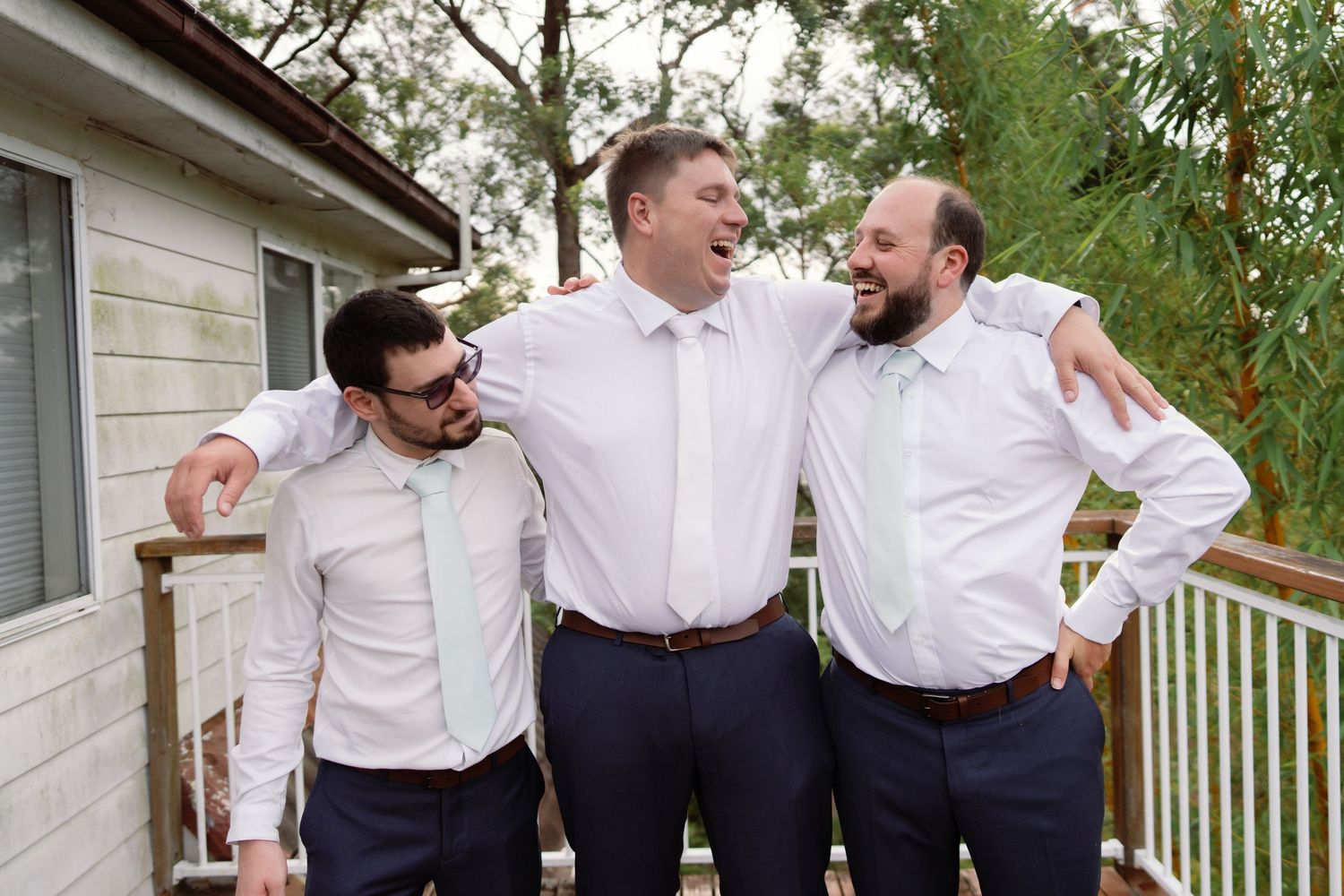 Groomsmen in white shirts and navy pants share a candid laughing moment together outdoors.