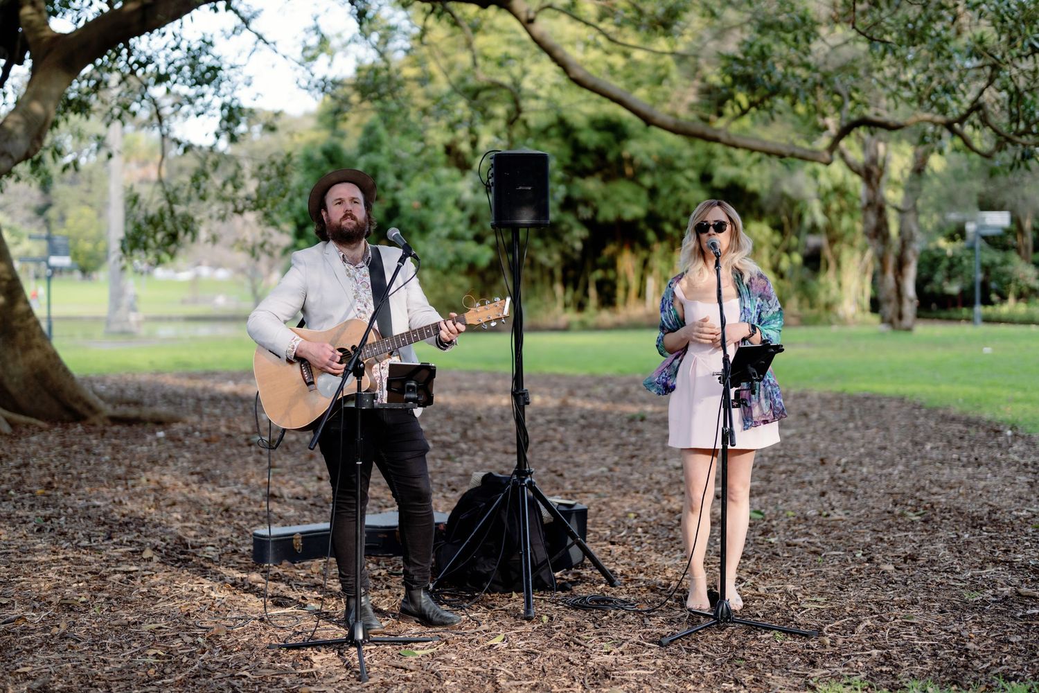 A man plays acoustic guitar while a woman sings beside a speaker stand outdoors in a park setting.