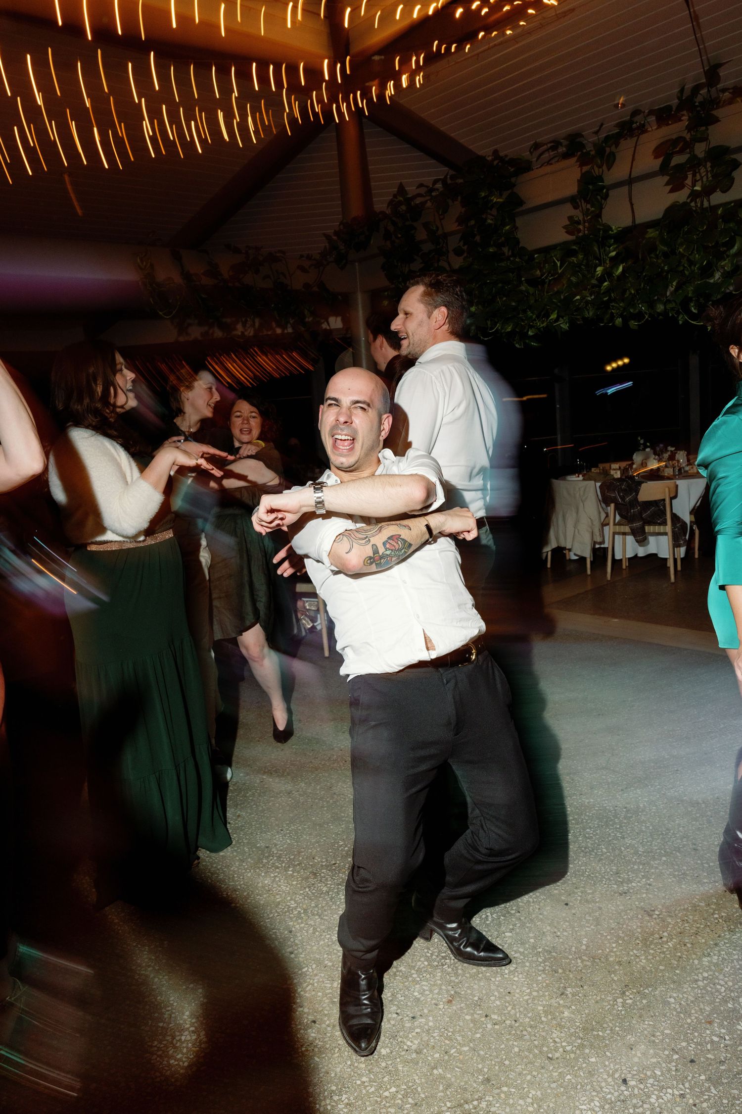 Guest energetically dances at a wedding reception under warm string lights in a decorated venue.