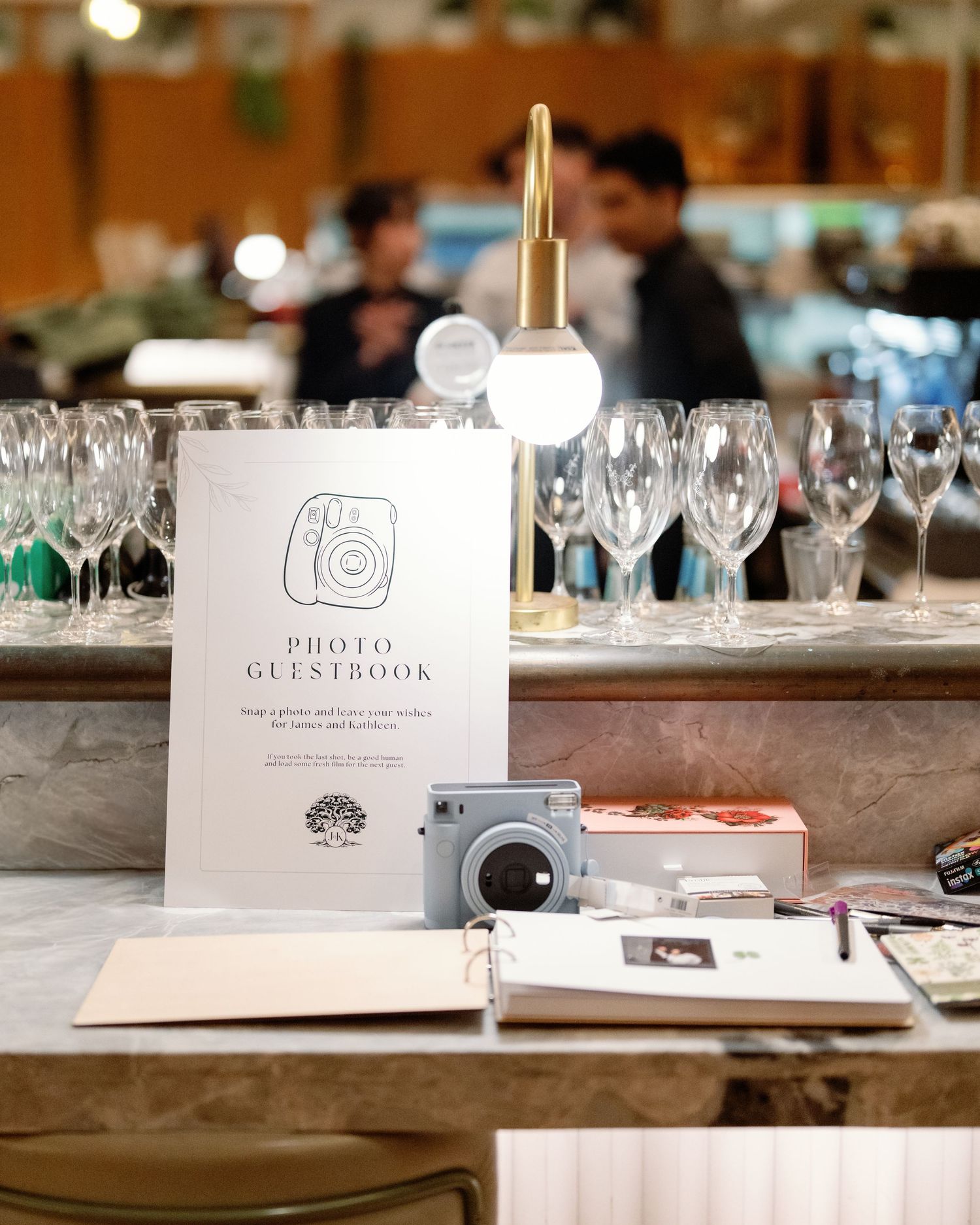 Wedding photo guestbook station with Instax camera, wine glasses, and lamp on marble bar.