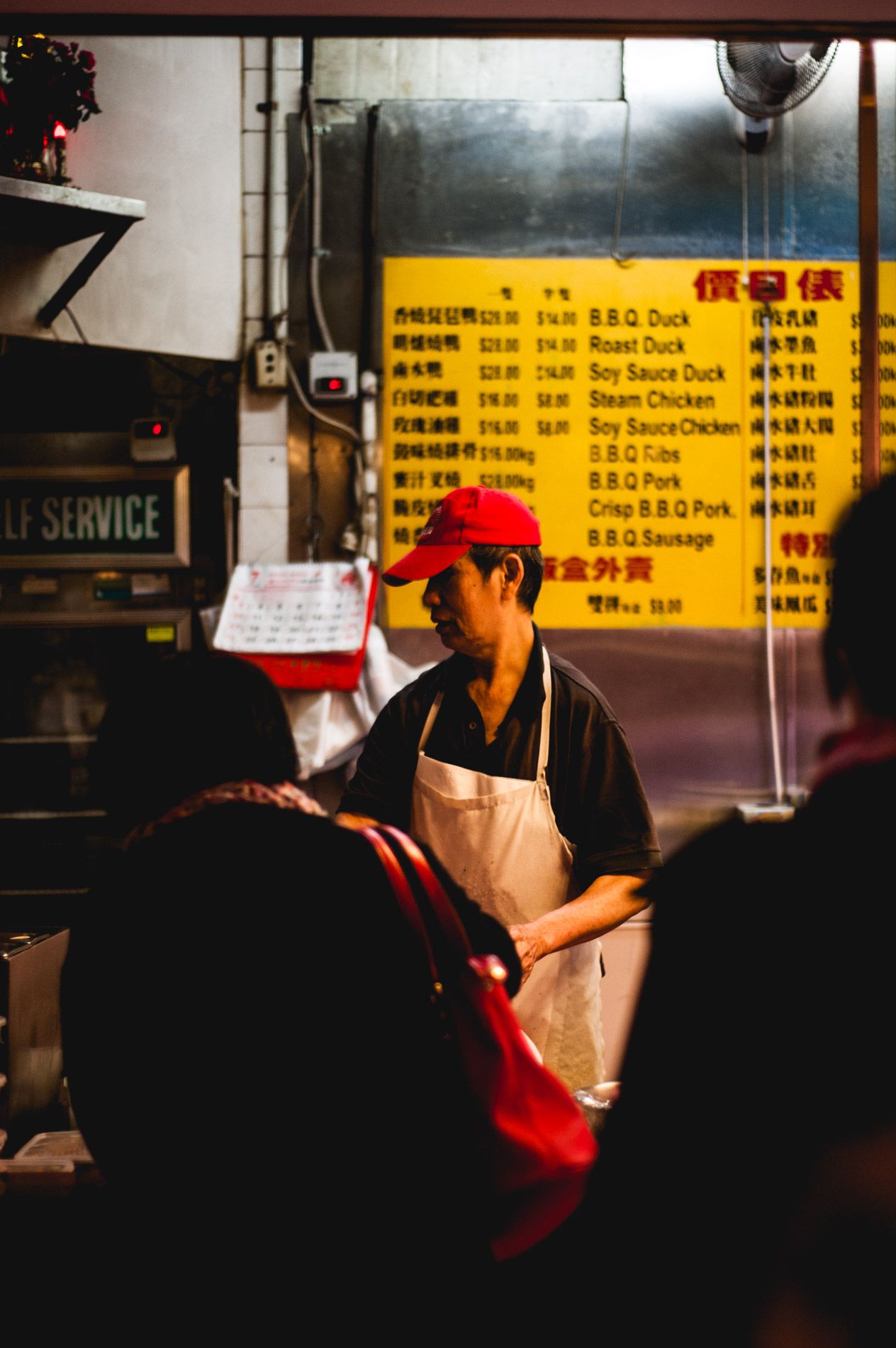 Restaurant worker in apron and cap stands in front of yellow menu board with Chinese text.