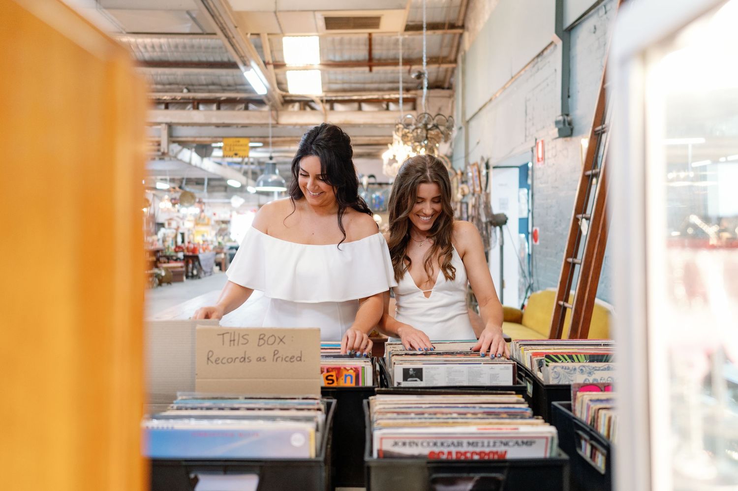 Two friends browse through vinyl records together in a music shop with orange walls and industrial ceiling.