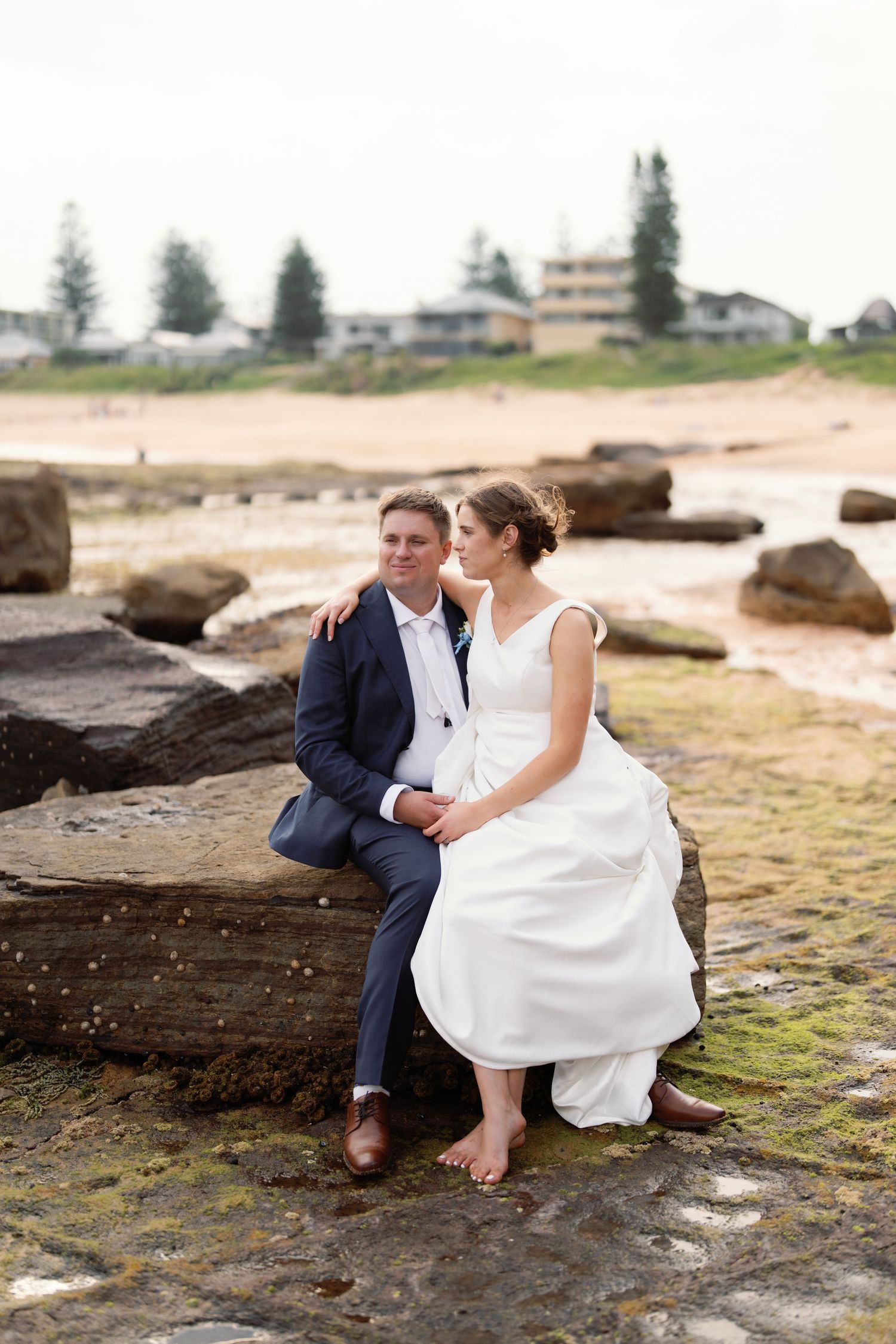 Wedding couple sitting on coastal rocks with beach and buildings in background.