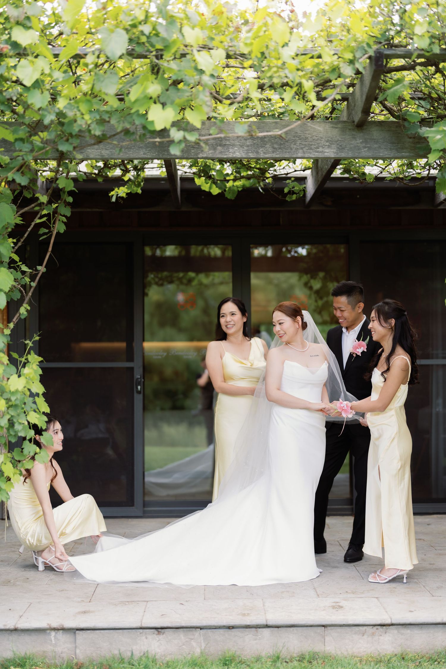Wedding party helping with dress preparations under vine-covered outdoor pavilion.