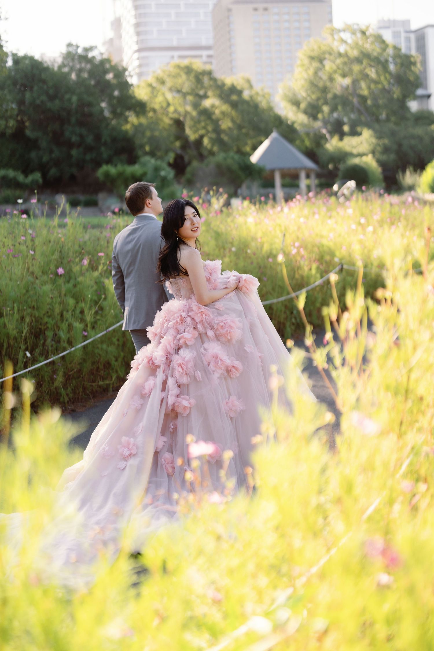 A couple in wedding attire poses in a sunlit meadow with tall grass and flowers at golden hour.