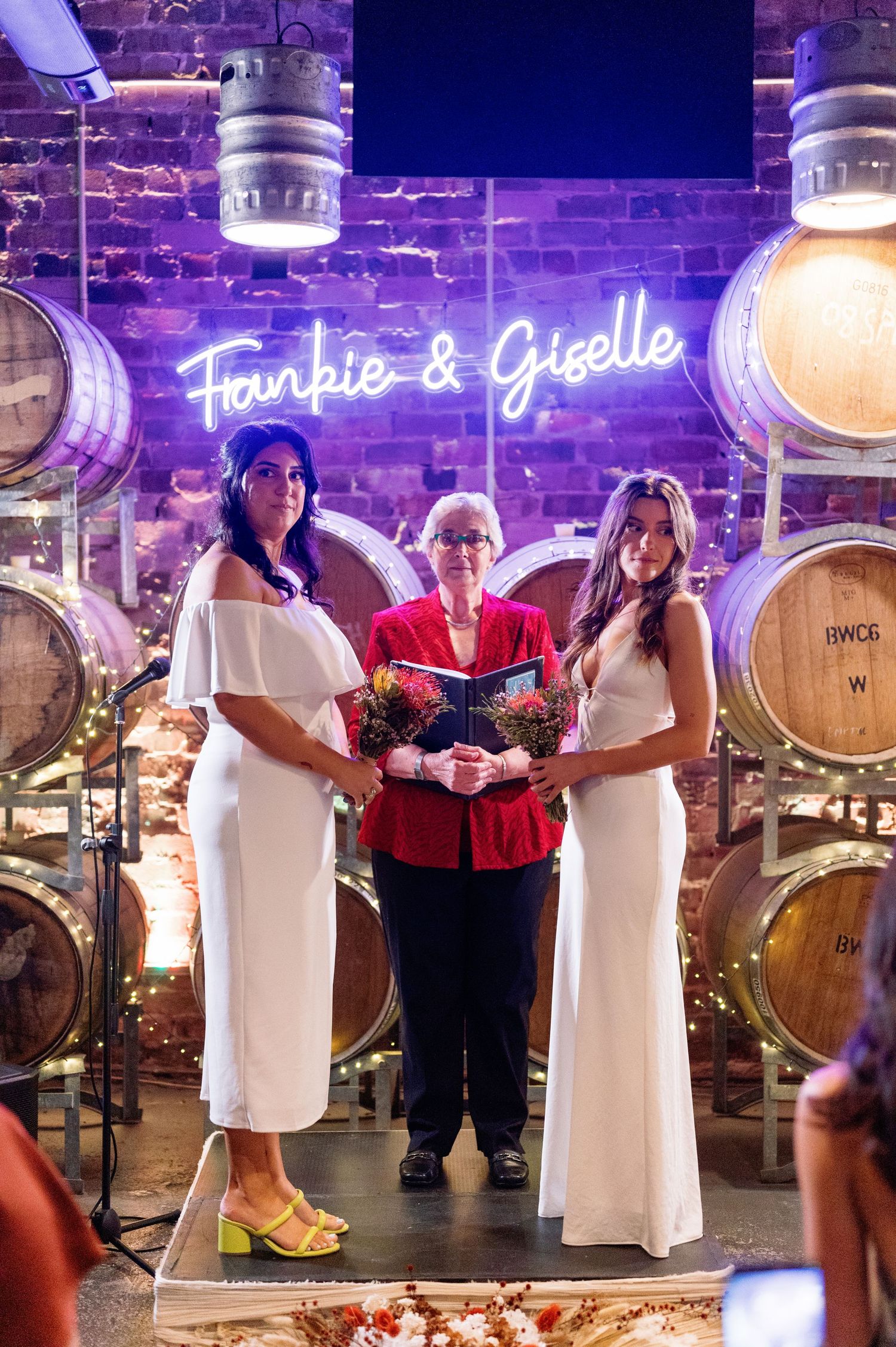 People in white and red attire gather in front of wooden wine barrels under purple lighting.