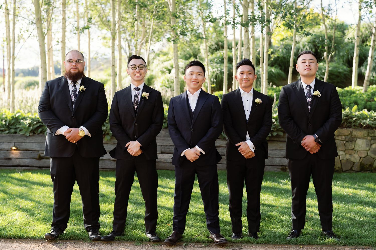 Five groomsmen in dark suits stand in formation on grass with trees in background.