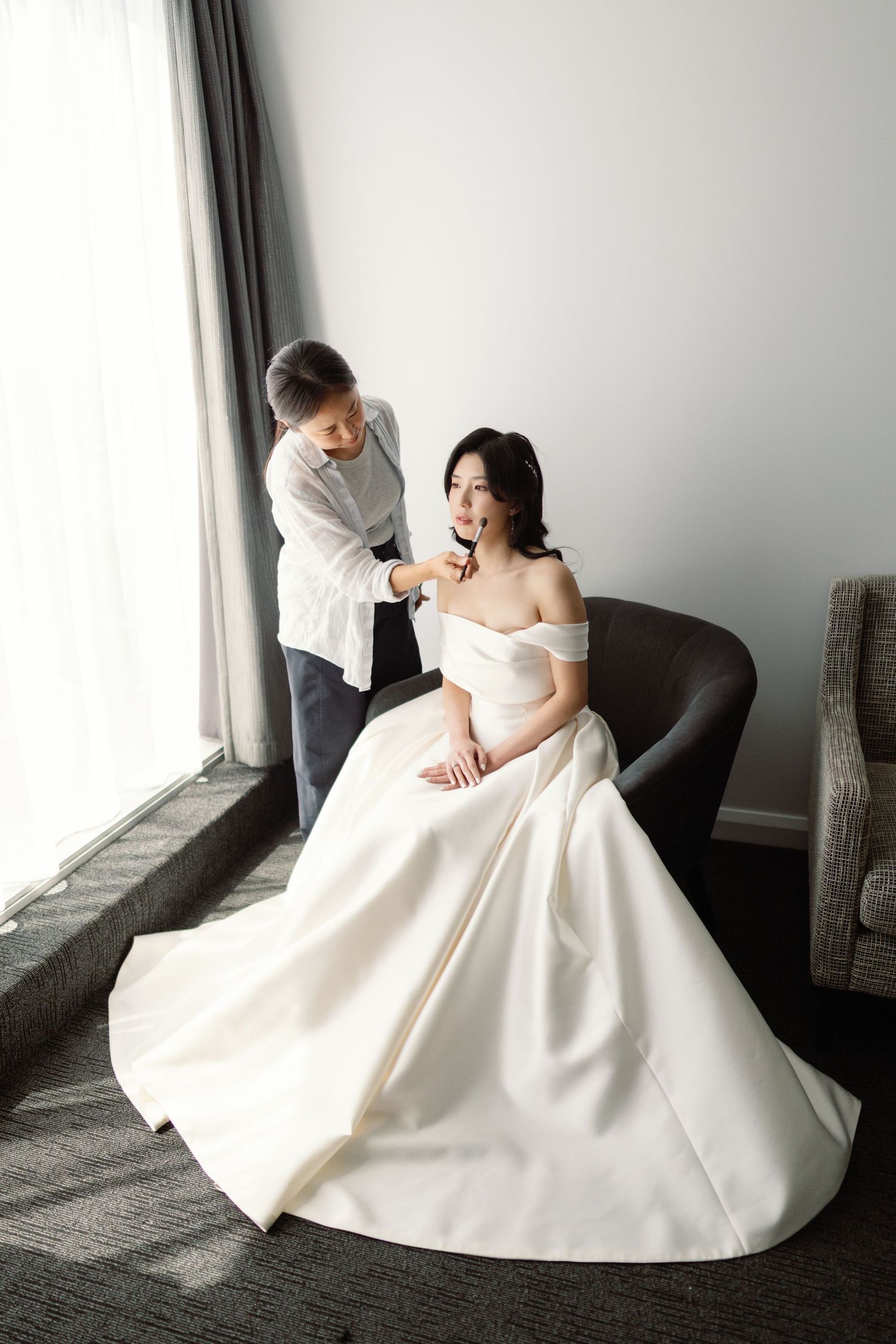 Elegant bride in off-shoulder ballgown poses gracefully in minimalist room with natural lighting.