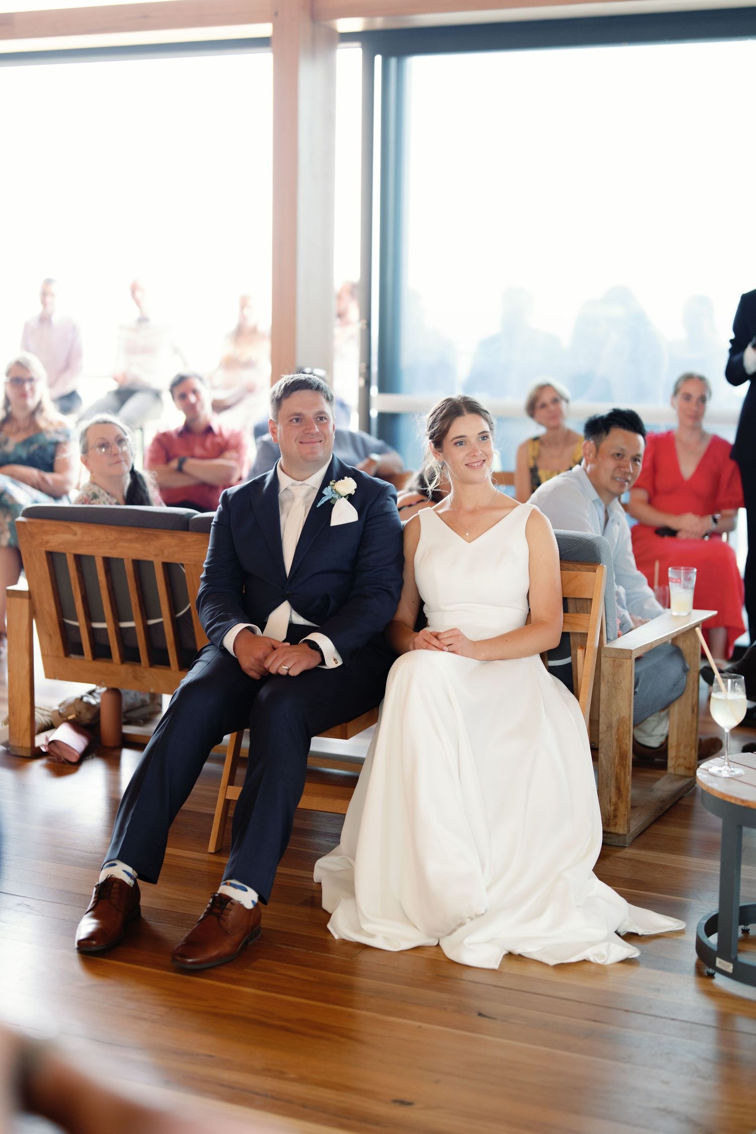 Wedding guests seated on wooden benches in a bright venue watching a ceremony unfold.
