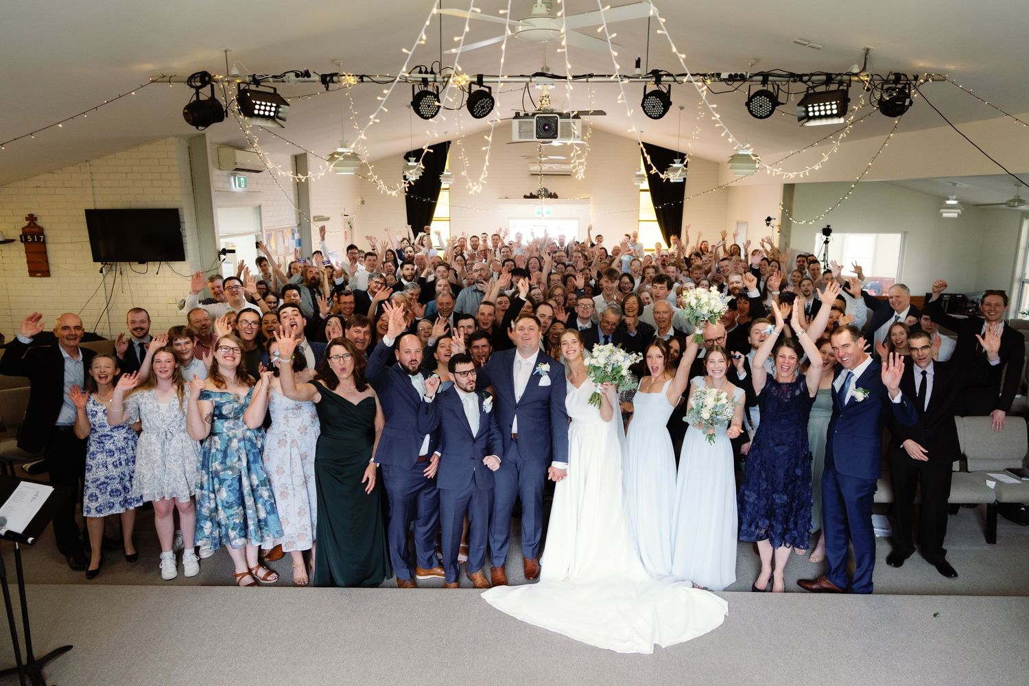 Large wedding reception hall filled with guests celebrating and raising their hands.