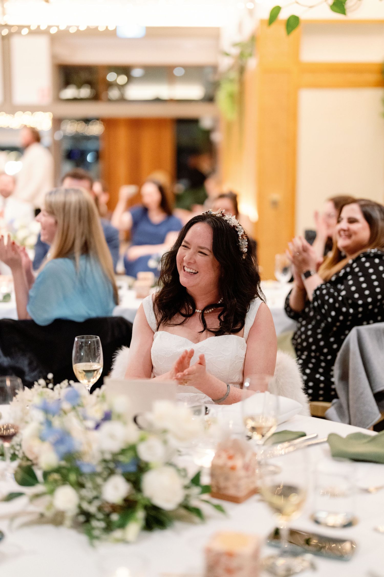 Bride laughing and clapping at wedding reception table surrounded by floral centerpieces and celebrating guests.