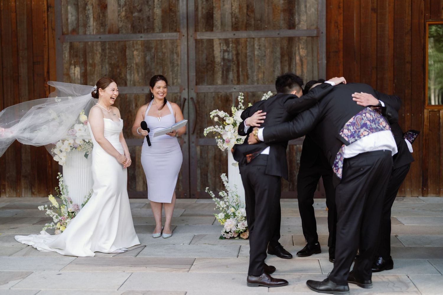 Wedding party posing for creative photos against rustic wooden barn backdrop.