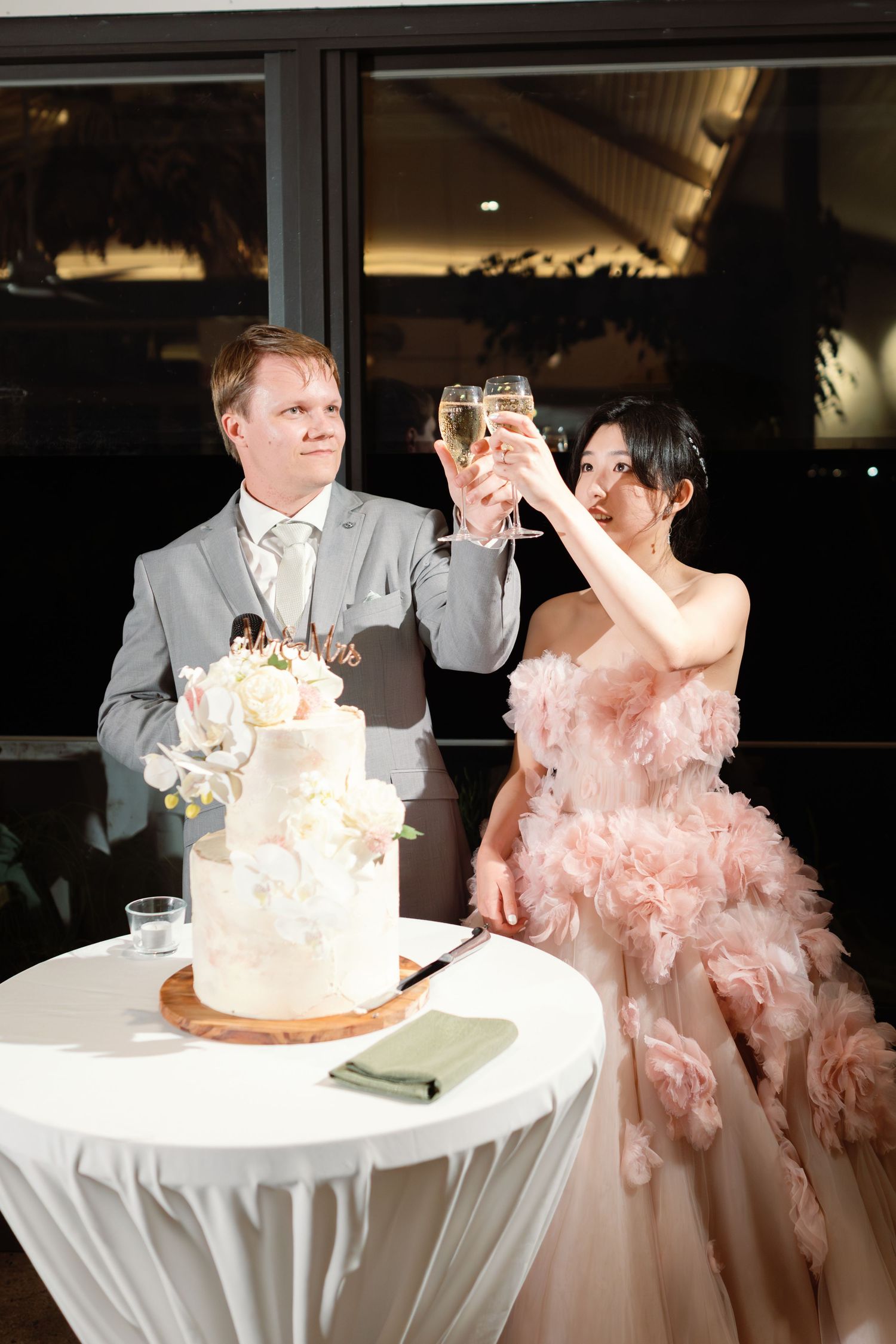A couple cutting their elegant white wedding cake with floral decorations while the bride wears a pink ruffled gown.