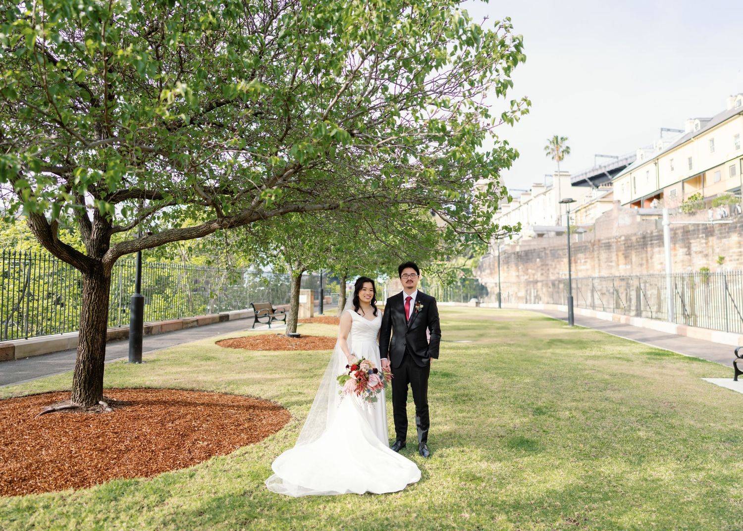 A wedding couple walks together on a lush green lawn with trees and buildings in the background.
