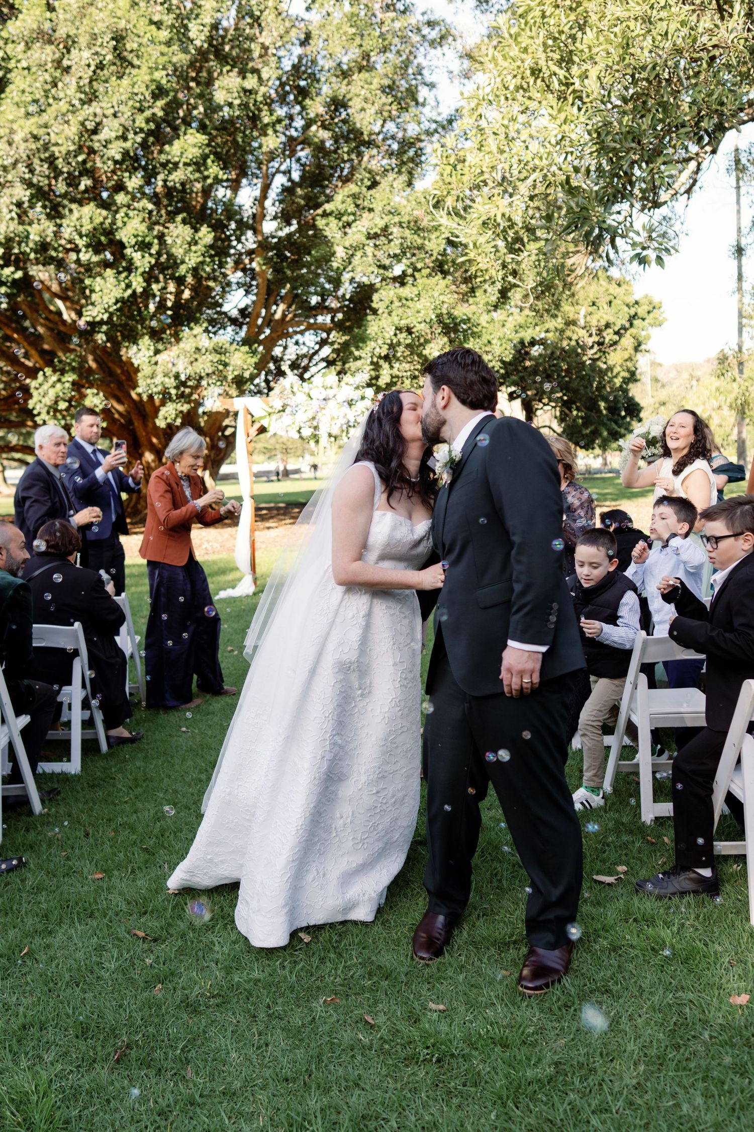 Bride and groom share a romantic kiss while guests blow bubbles during outdoor wedding ceremony recessional.