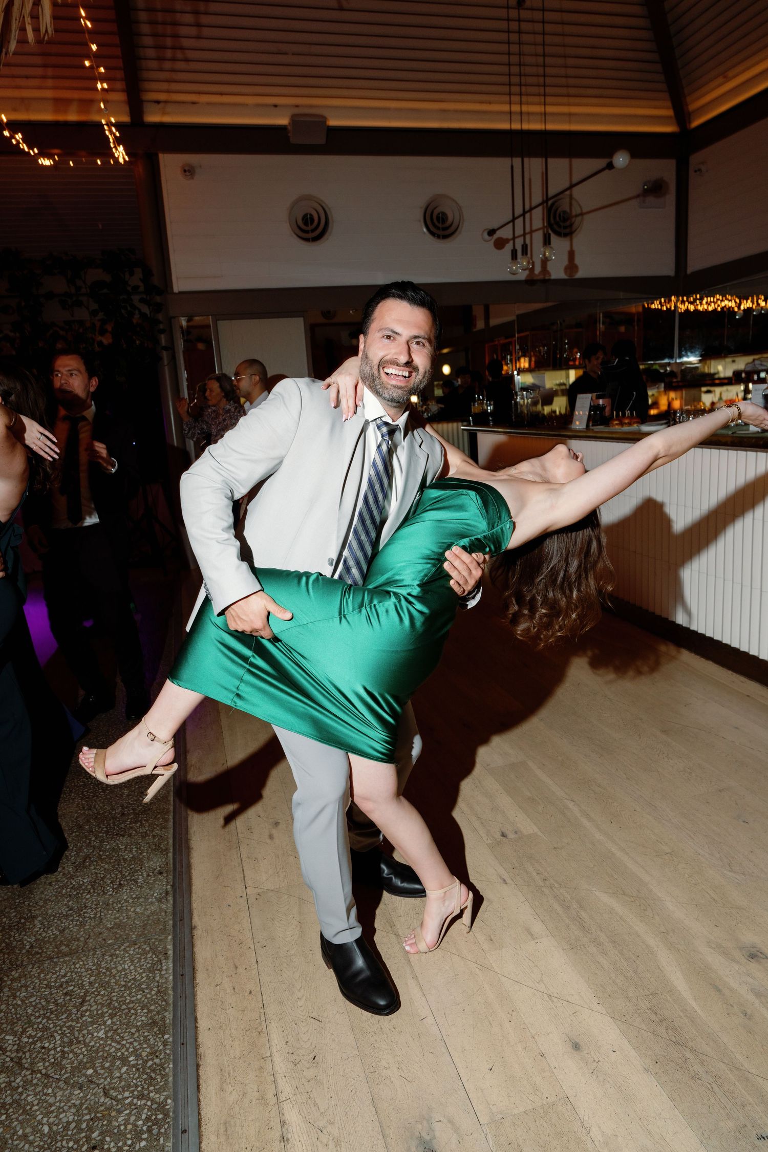 Man in gray suit lifts woman in green dress while dancing at an elegant indoor wedding reception.