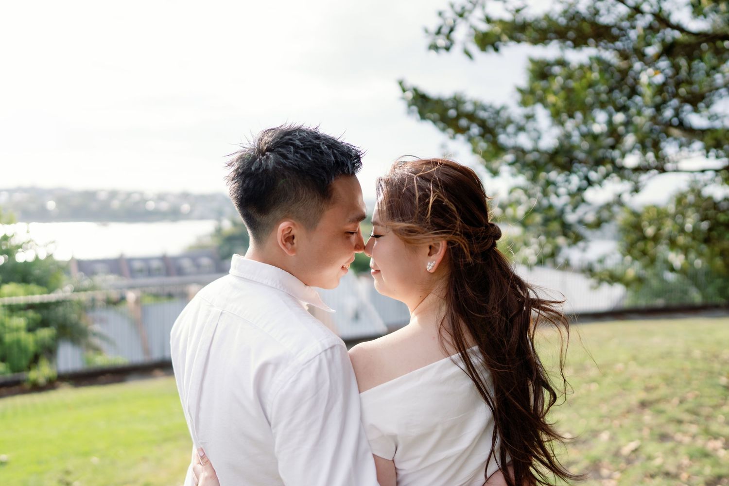 A couple in white outfits shares a romantic moment on a grassy hilltop overlooking a scenic coastal view.