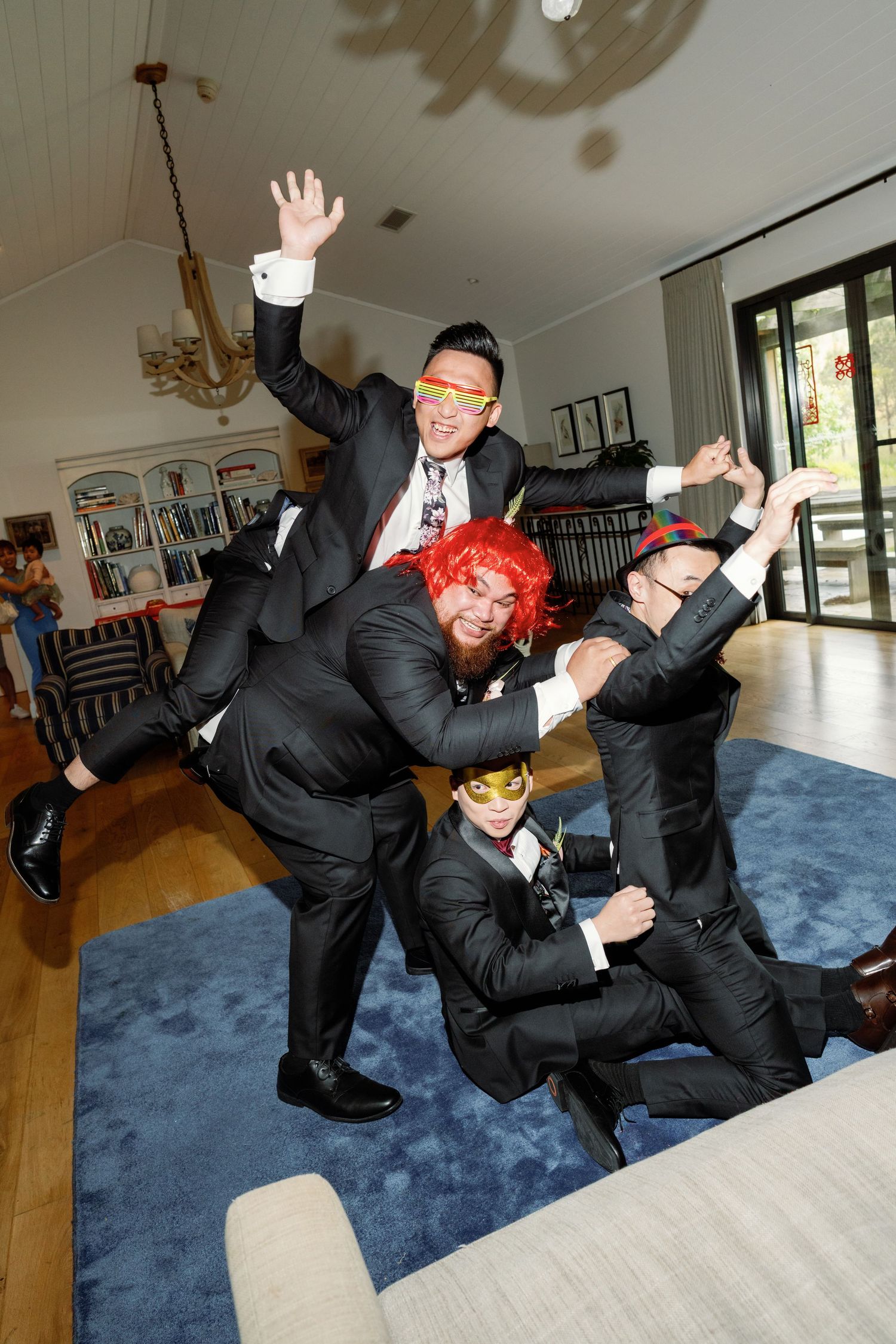 Groomsmen in black suits perform a fun jumping pose sequence on a blue carpet indoors.