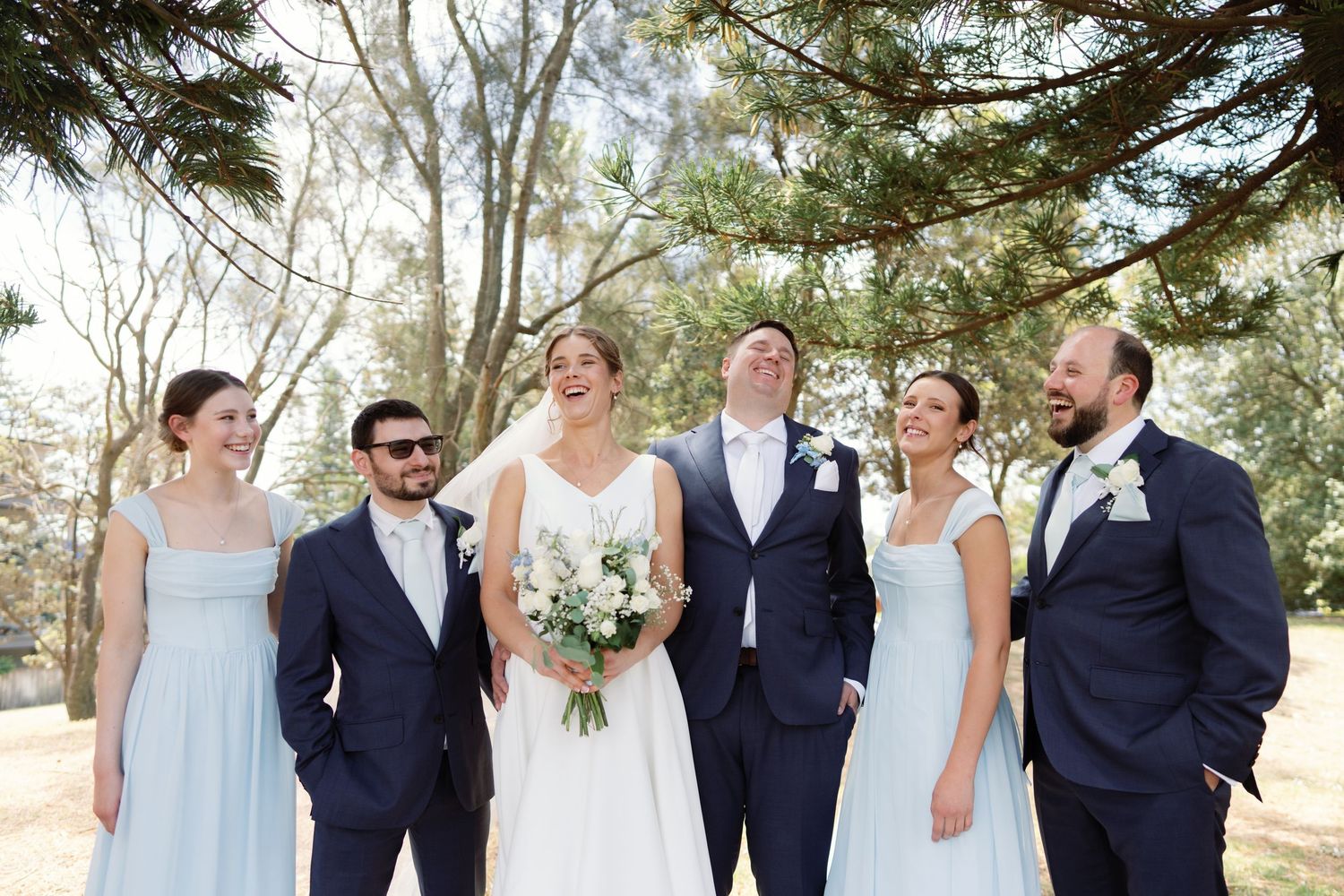 Wedding party in navy and light blue attire posing together outdoors among trees.