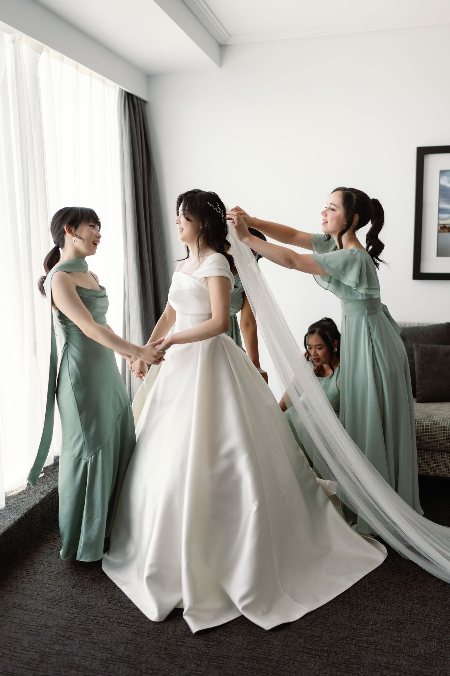 Bridesmaids in sage green dresses help adjust a white wedding gown during pre-ceremony preparations.