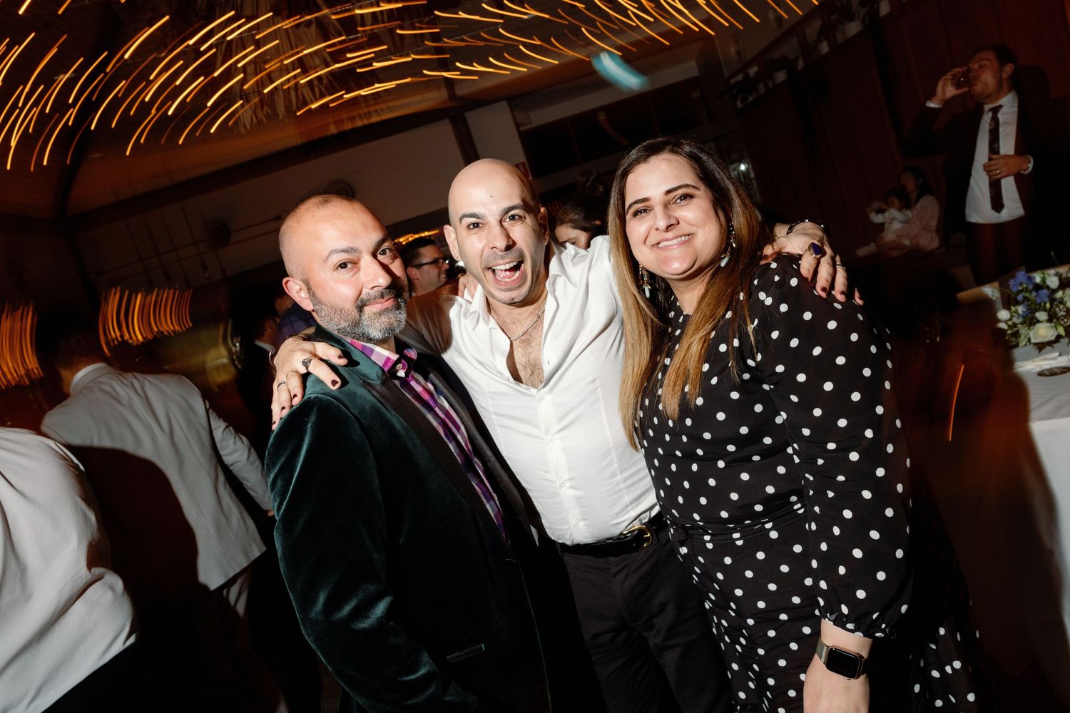 Three friends smiling and posing together at a lively indoor party, woman wearing polka dot dress.
