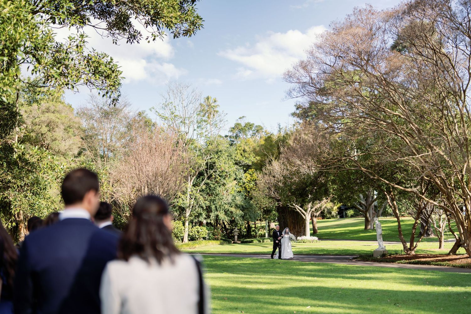 Outdoor wedding ceremony on lush green lawn surrounded by tall trees under a bright blue sky.
