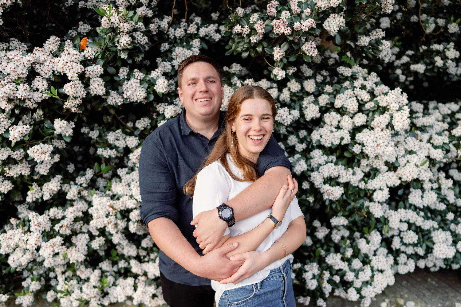 Smiling couple embracing in front of a lush white flowering bush in an outdoor garden setting.
