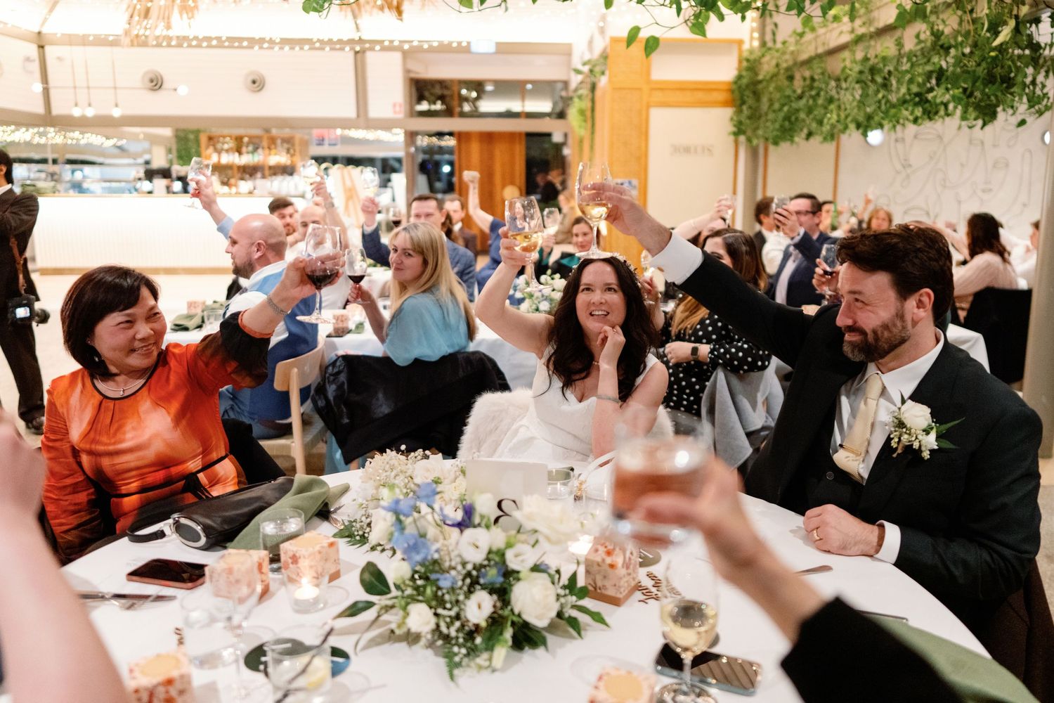 Wedding guests raise glasses in a toast at a beautifully decorated reception table with floral centerpiece.