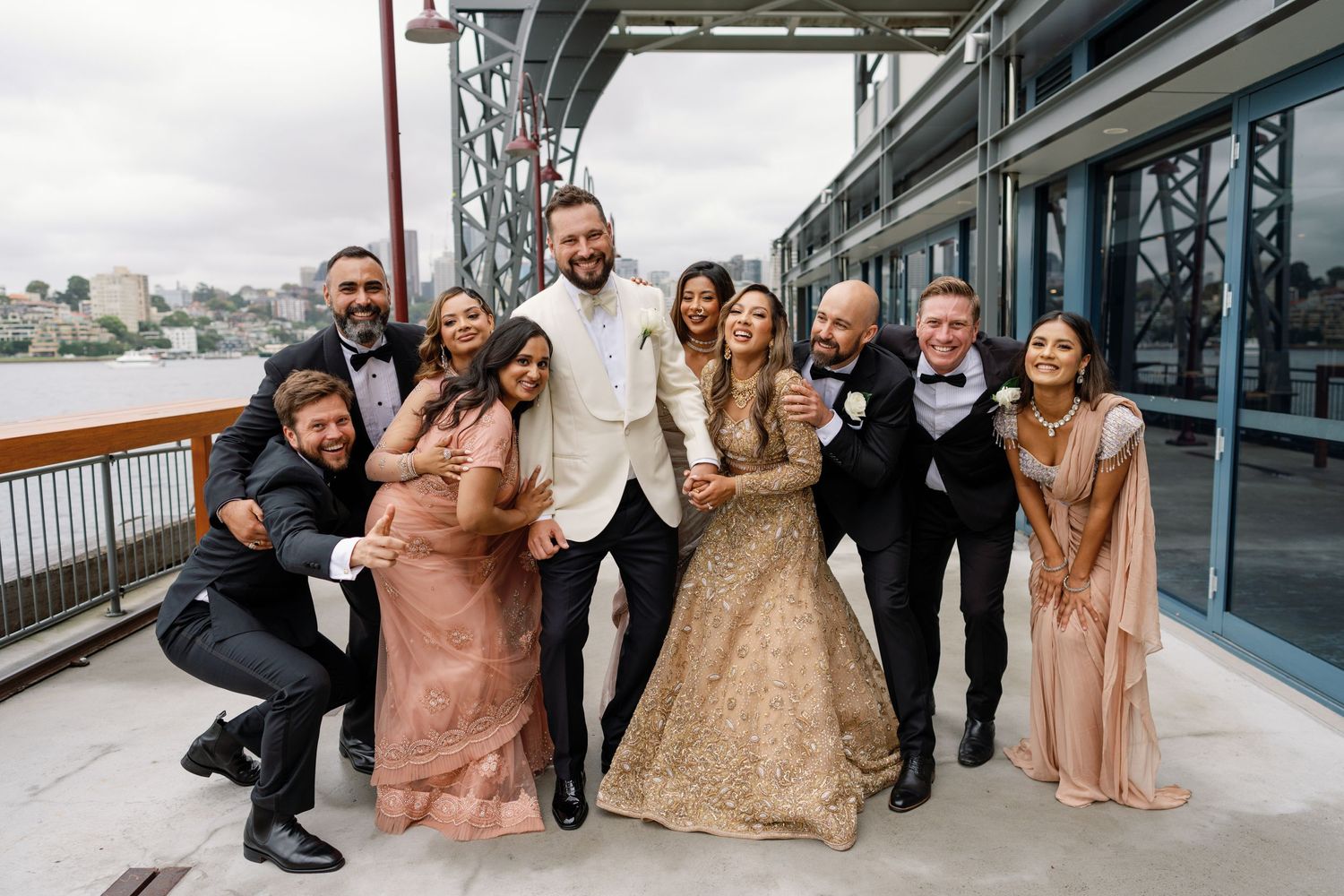 Wedding party in formal attire posing together on a waterfront deck.