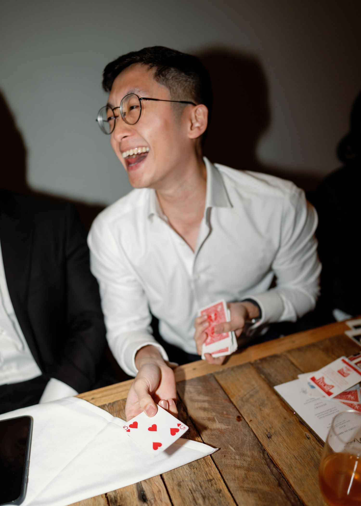 Person in white shirt laughing while playing cards at wooden table.