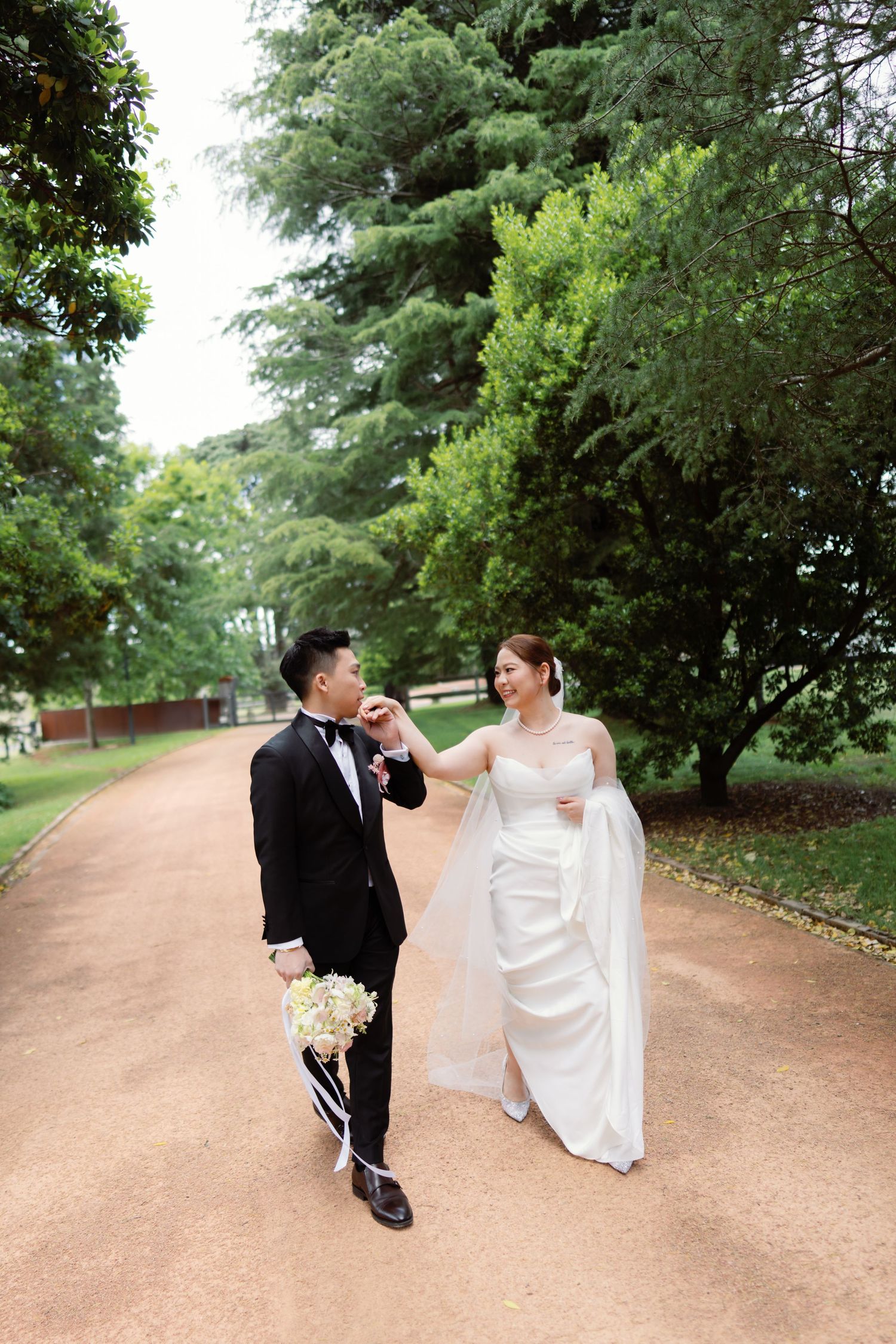 Newlyweds dance playfully on a dirt path surrounded by green trees.