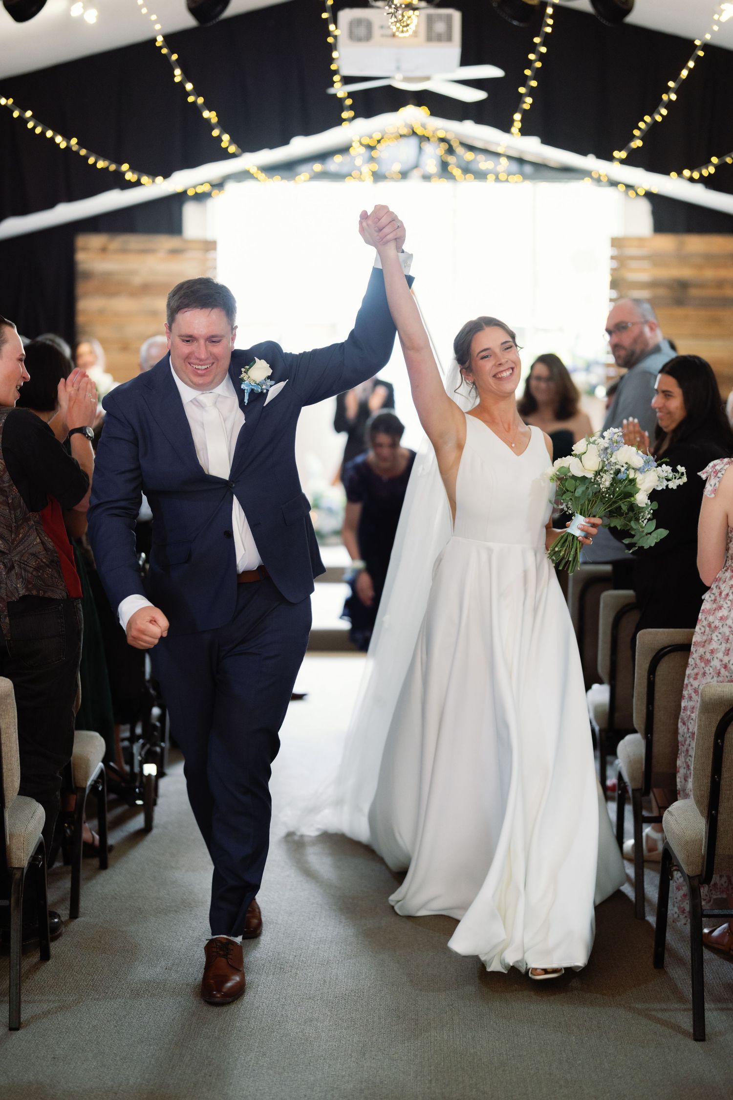 Wedding couple celebrating with joyful dance moves down the aisle under twinkling lights.