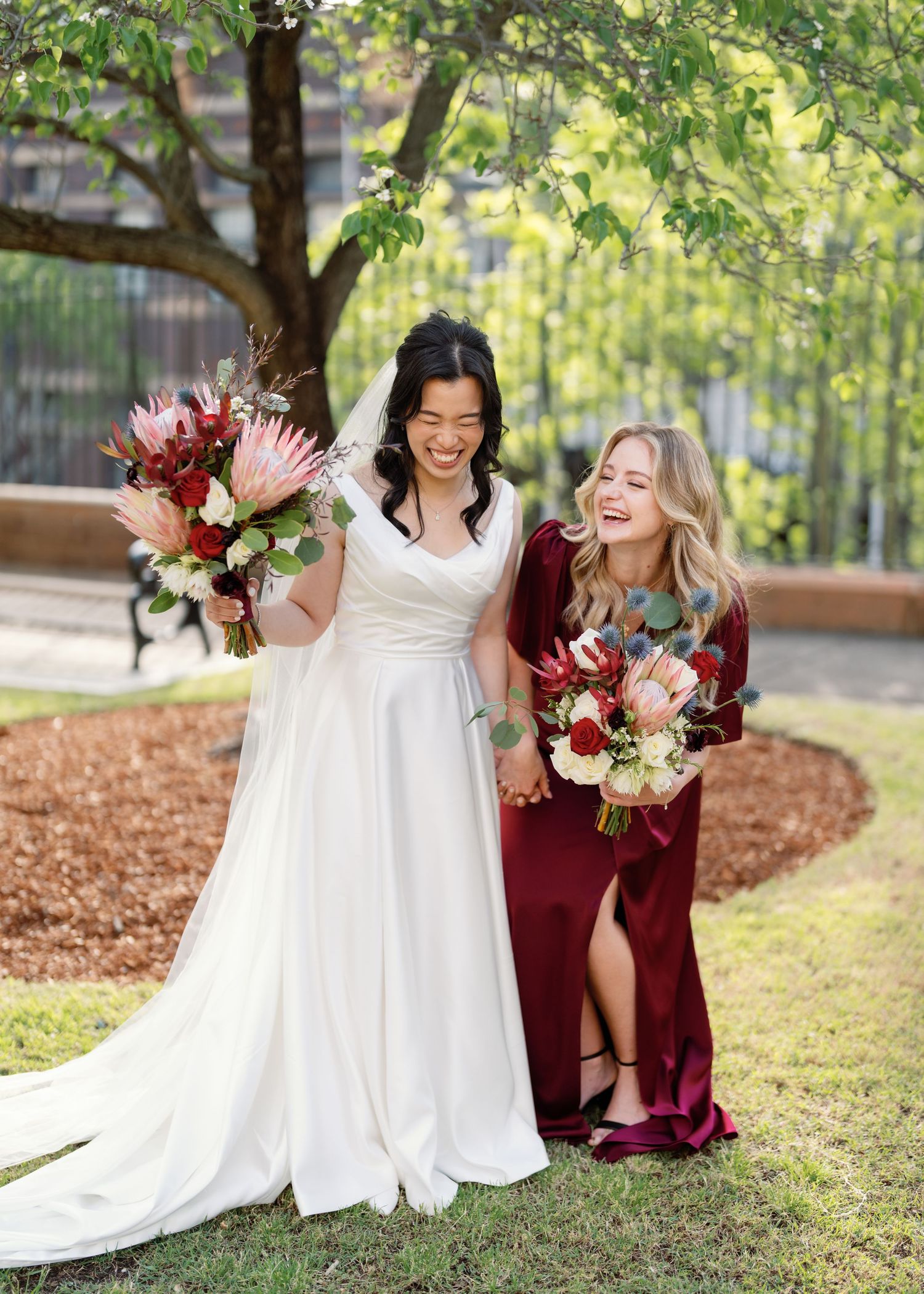 Wedding party in burgundy and white dresses laughing together on grass under trees.