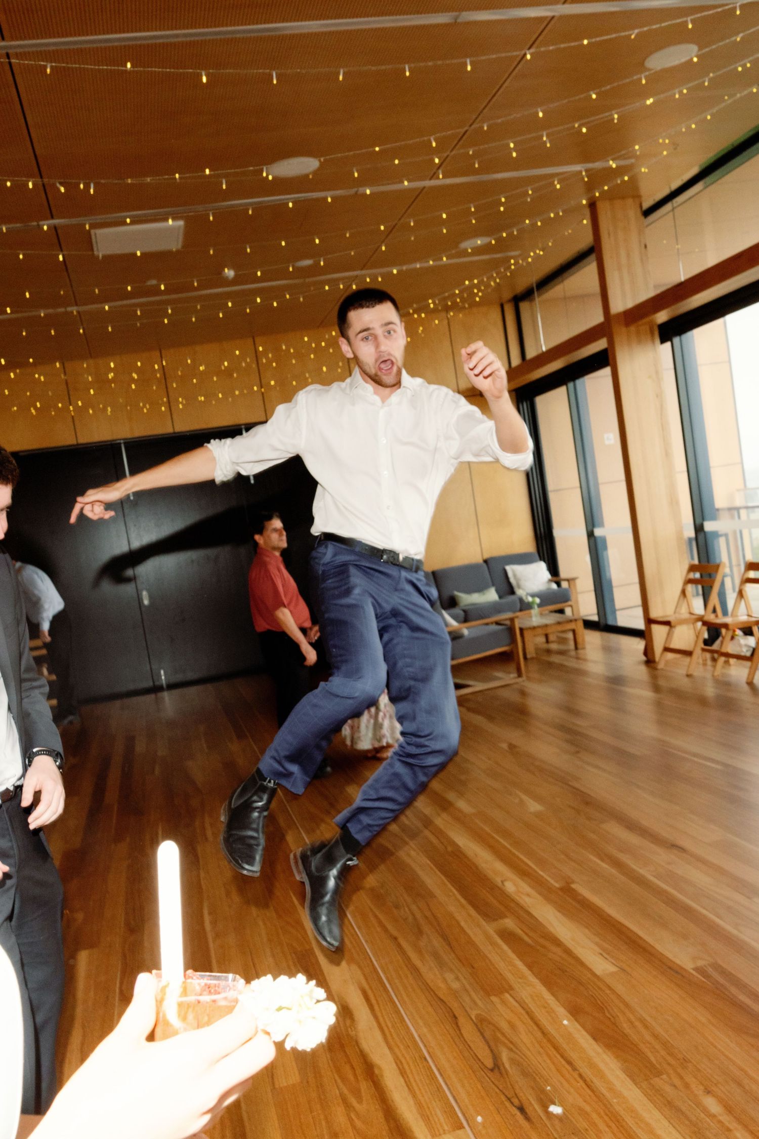 Wedding guests dancing energetically on wooden floor under string lights.