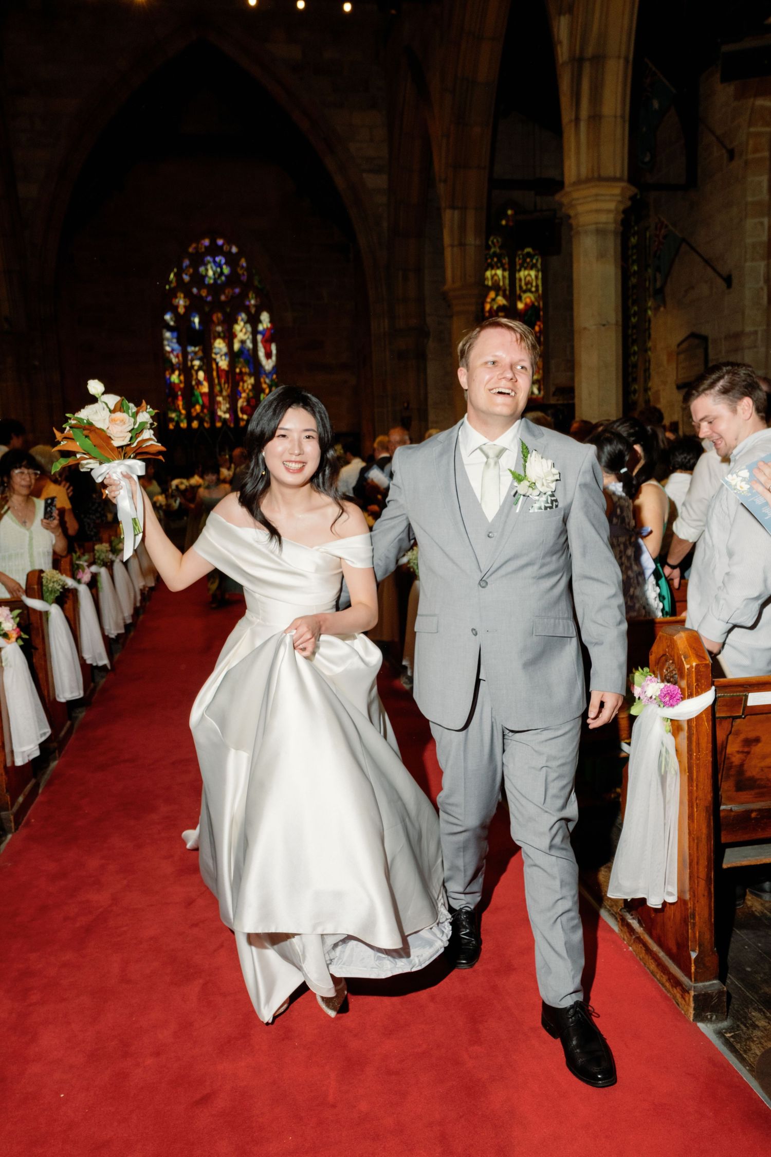 Wedding party walking down red carpet aisle in church wearing gray suits and white dress.