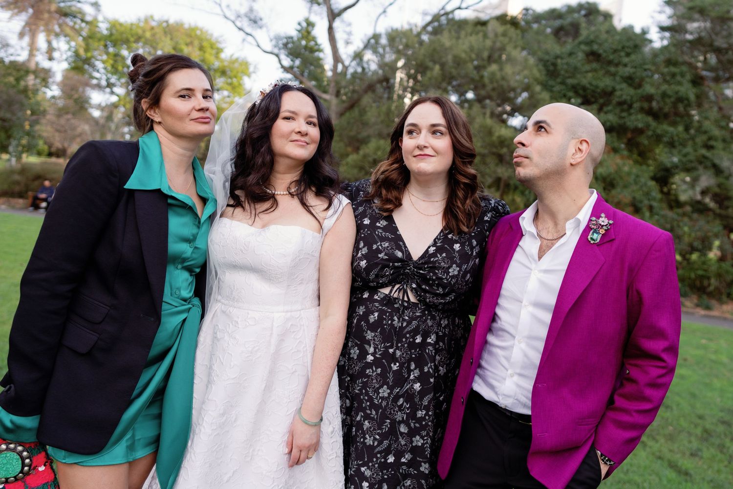 Four wedding guests pose outdoors, including a bride in white, wearing formal attire in vibrant colors.